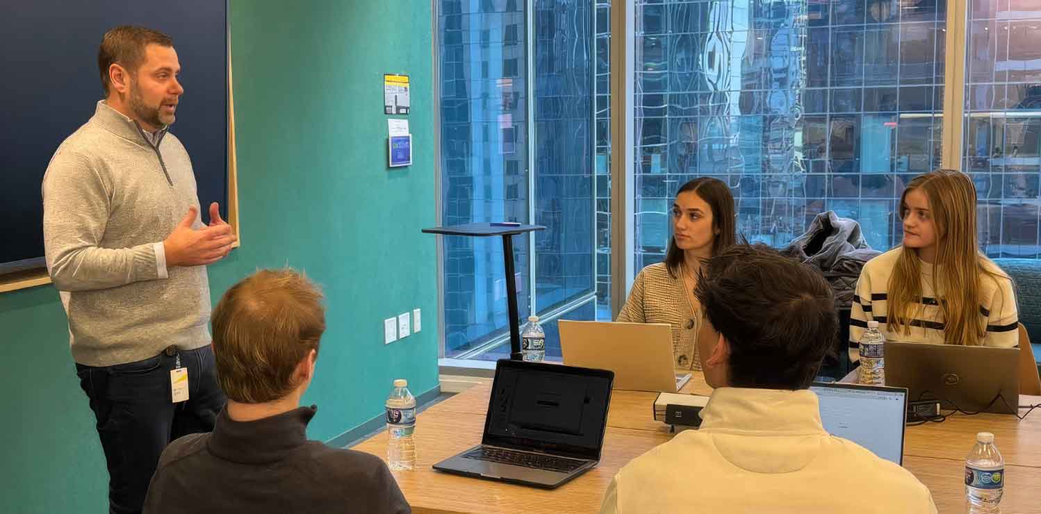 Man speaking to a small group in a meeting room while attendees listen with laptops open at a conference table, with tall city buildings visible through large windows.