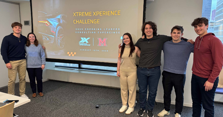 Group of six people standing in a conference room in front of a projected slide titled “XTREME XPERIENCE CHALLENGE,” with logos and a stylized car graphic.