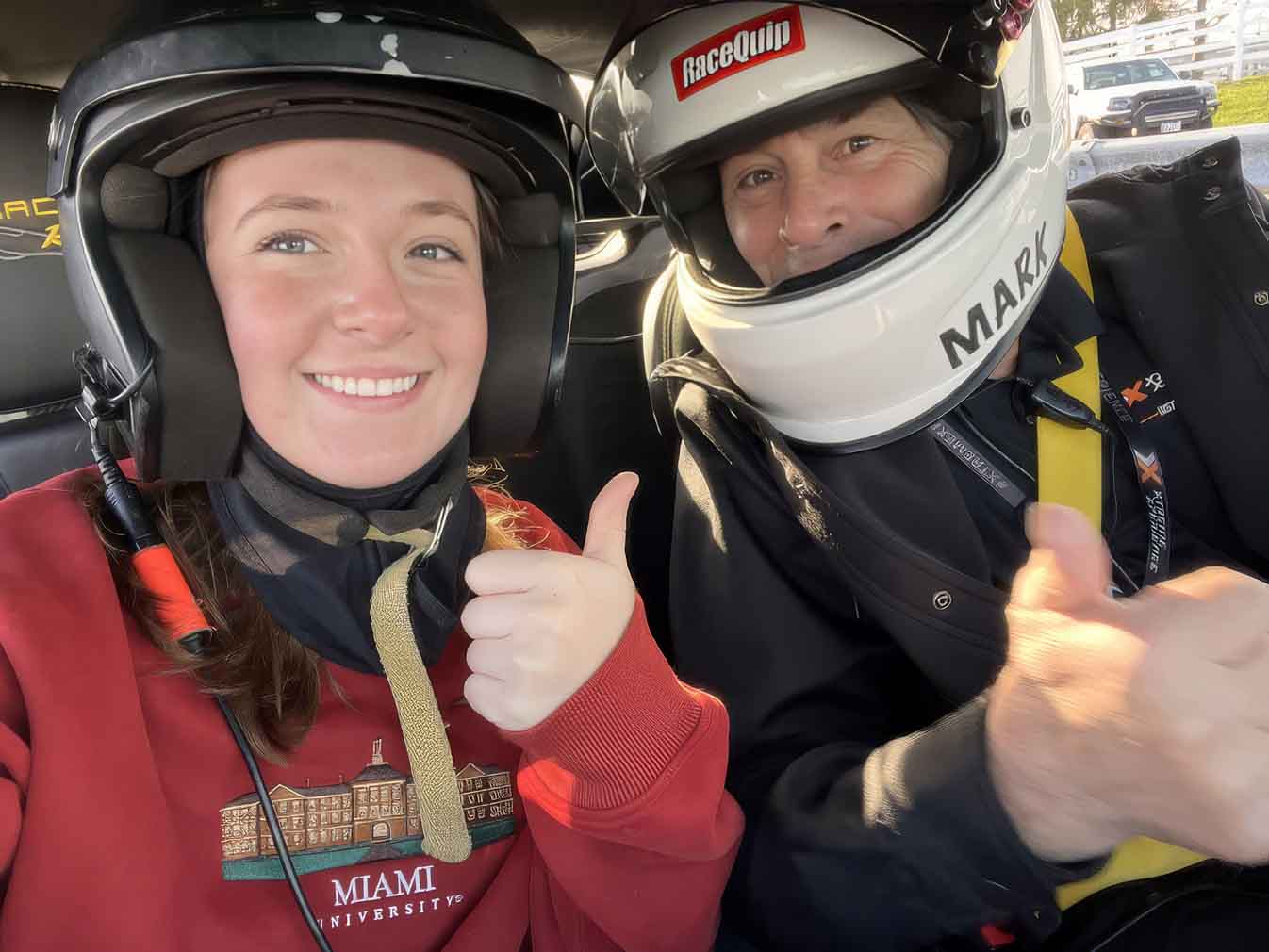 Two people wearing racing helmets sit in a car and give thumbs-up to the camera.