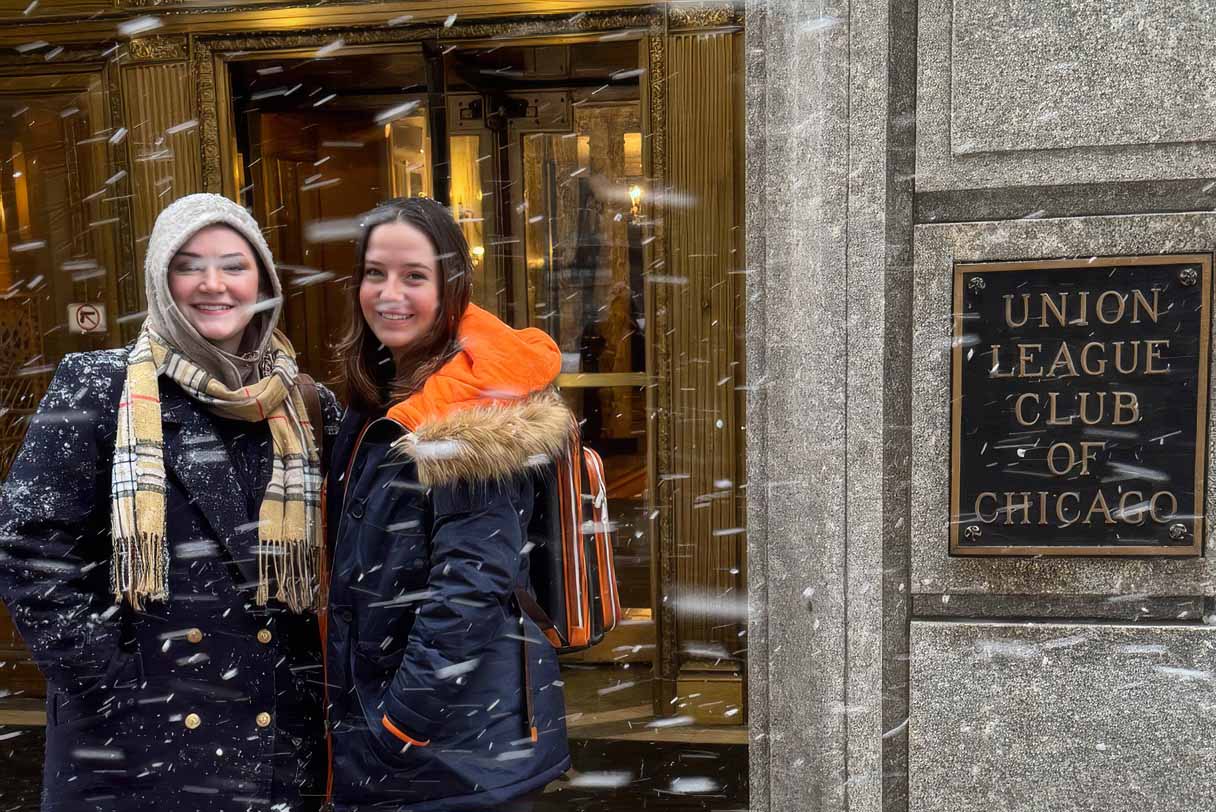 Two people in winter coats pose smiling in falling snow outside a building entrance beside a plaque reading “Union League Club of Chicago.”