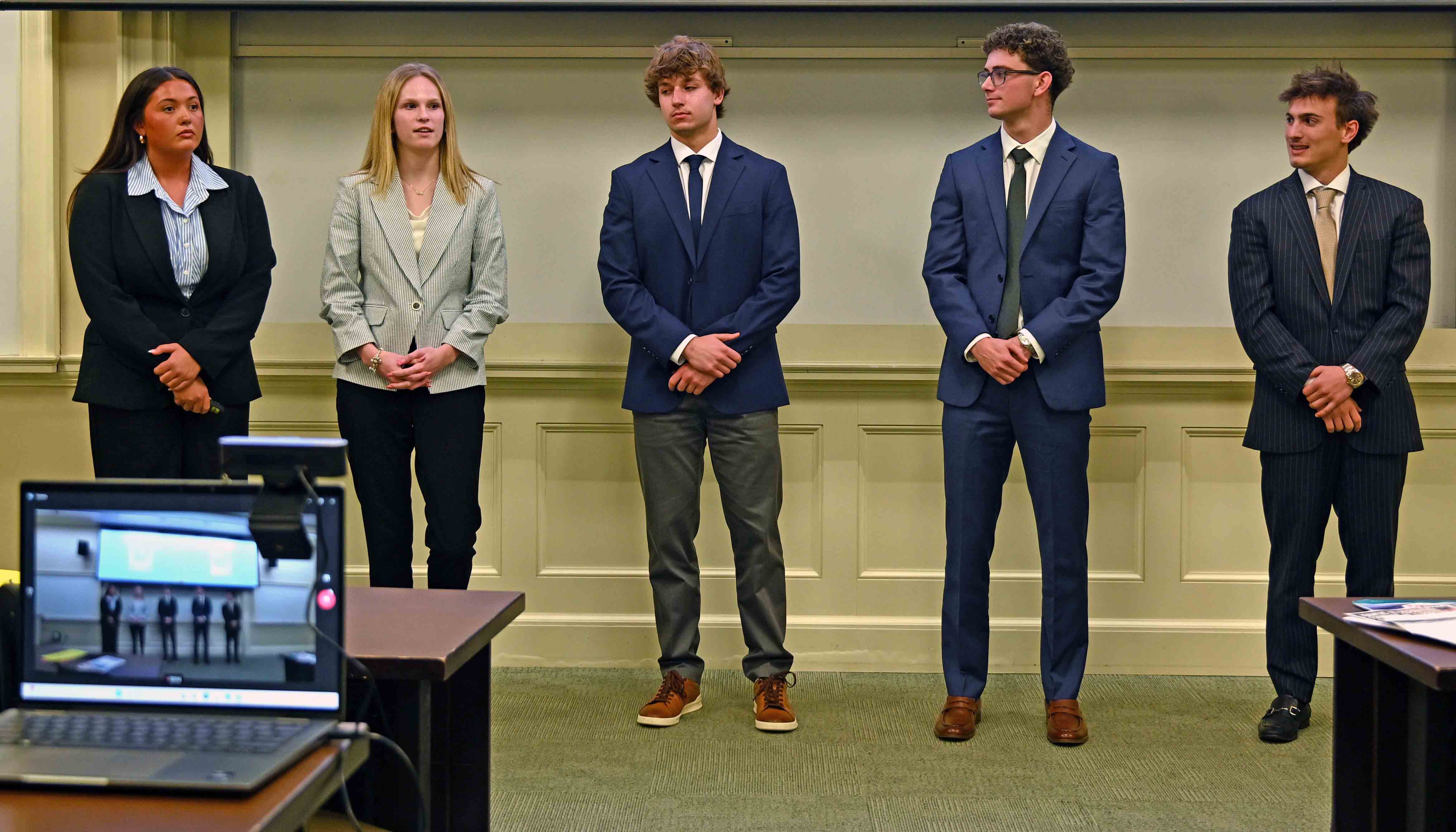 Five young adults in business attire stand in a line at the front of a classroom, as if presenting to an audience.