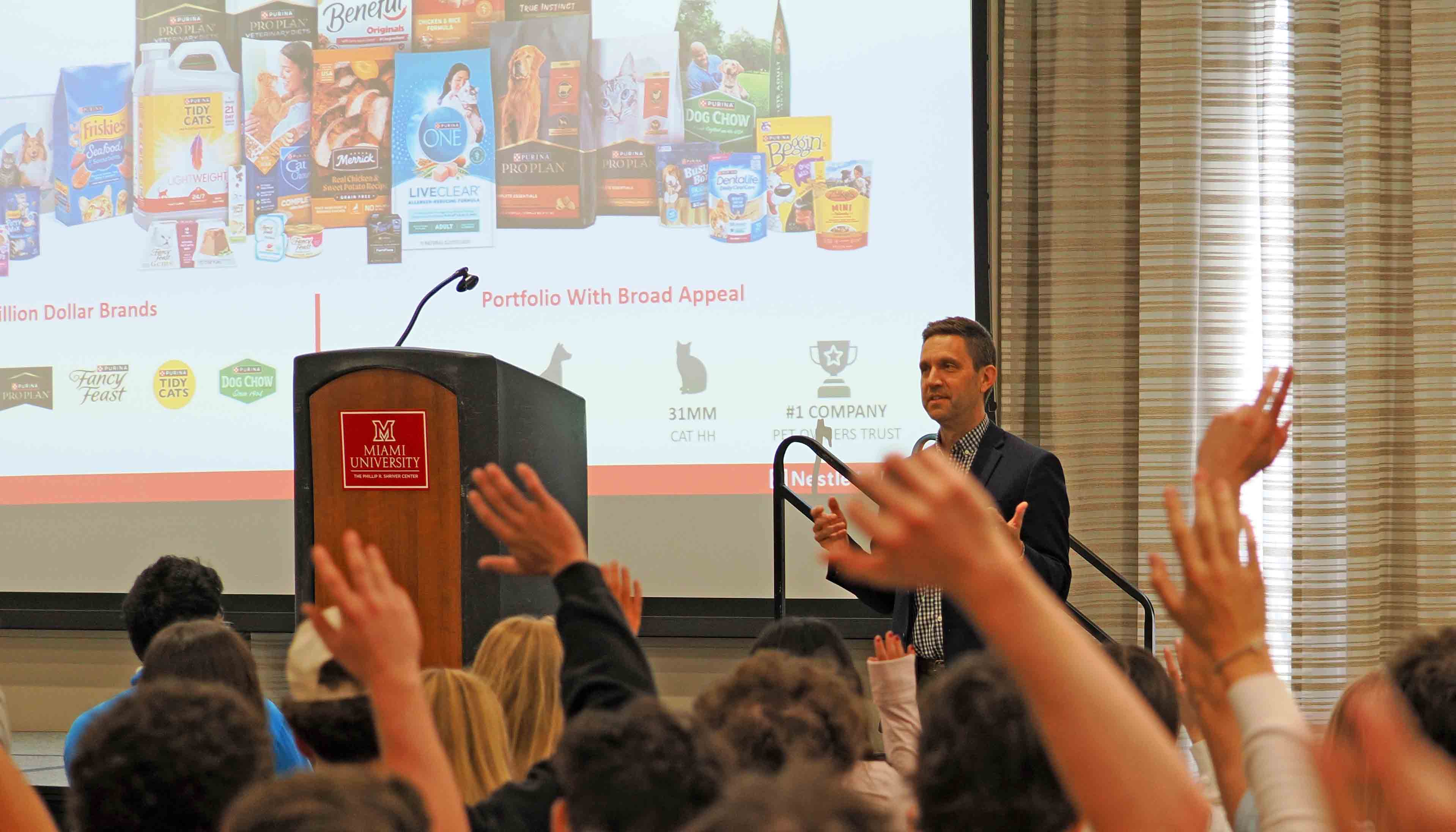 Guest speaker presenting to an audience in a lecture hall, with raised hands in the foreground and a slide of pet food products projected behind him.
