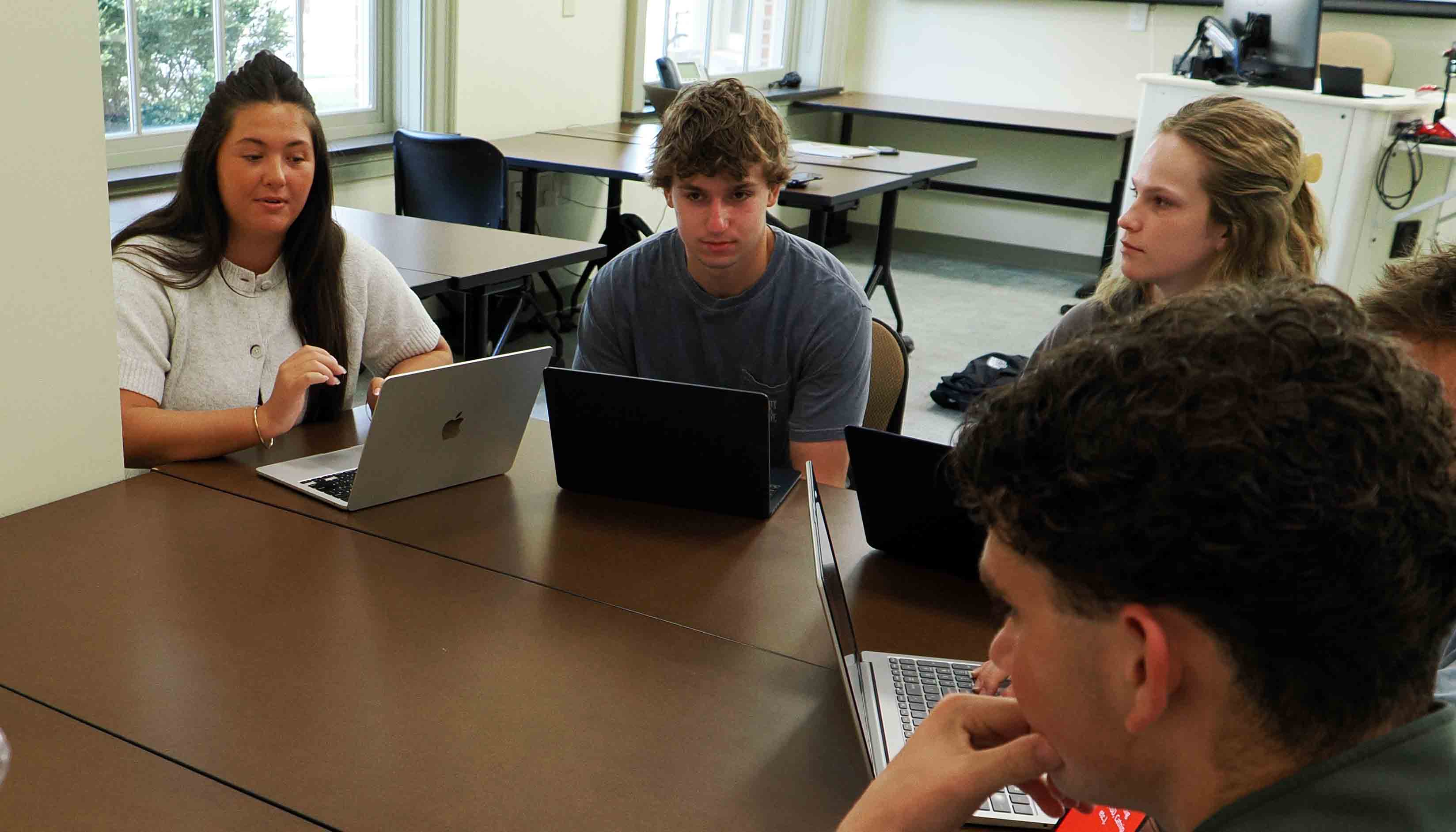 Three students sit around a conference table with laptops during a group meeting, with a career ambassador presentation displayed on a wall-mounted screen behind them.