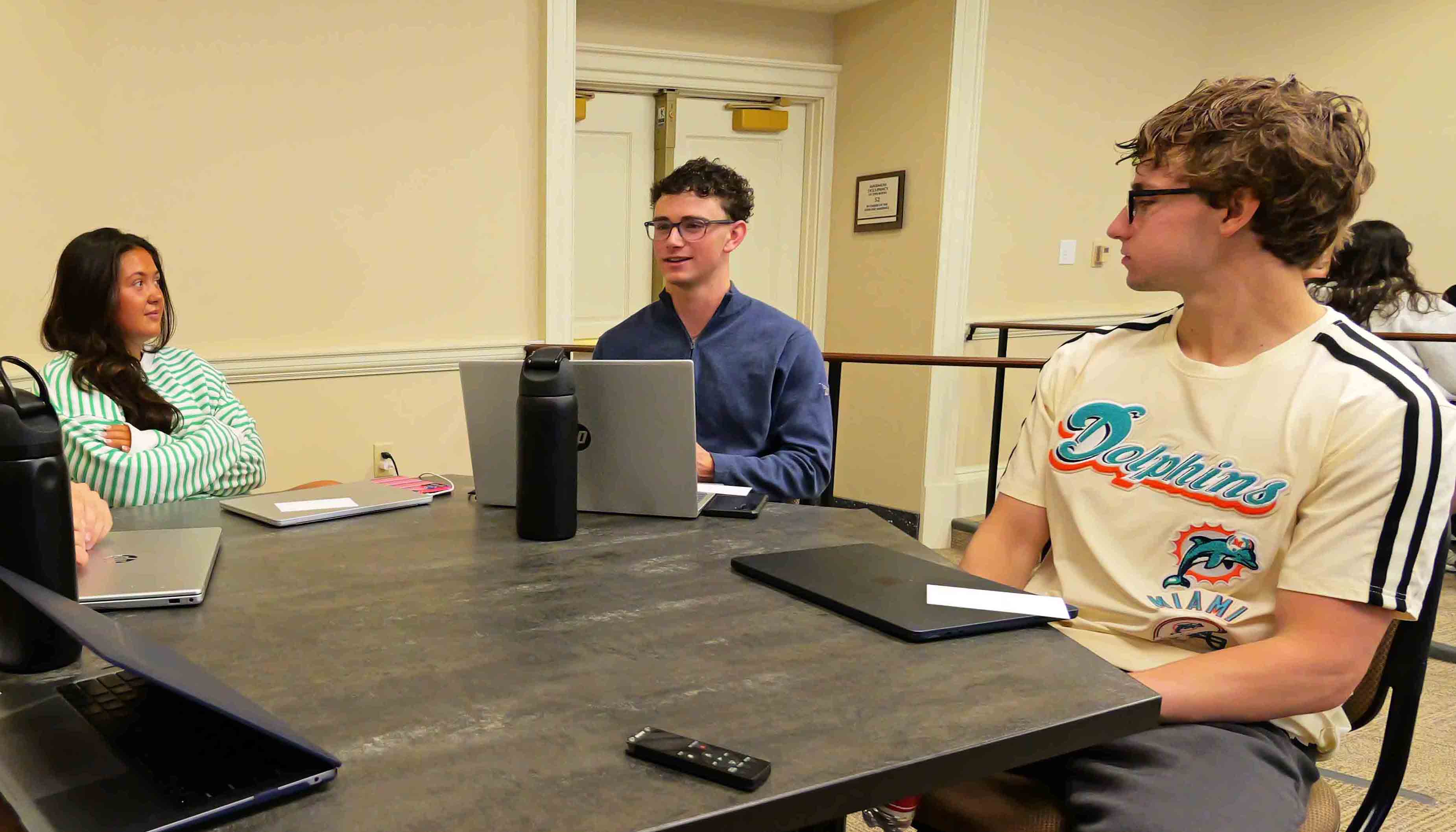 Students sit around a table with laptops, listening to one person speak during a small group meeting.