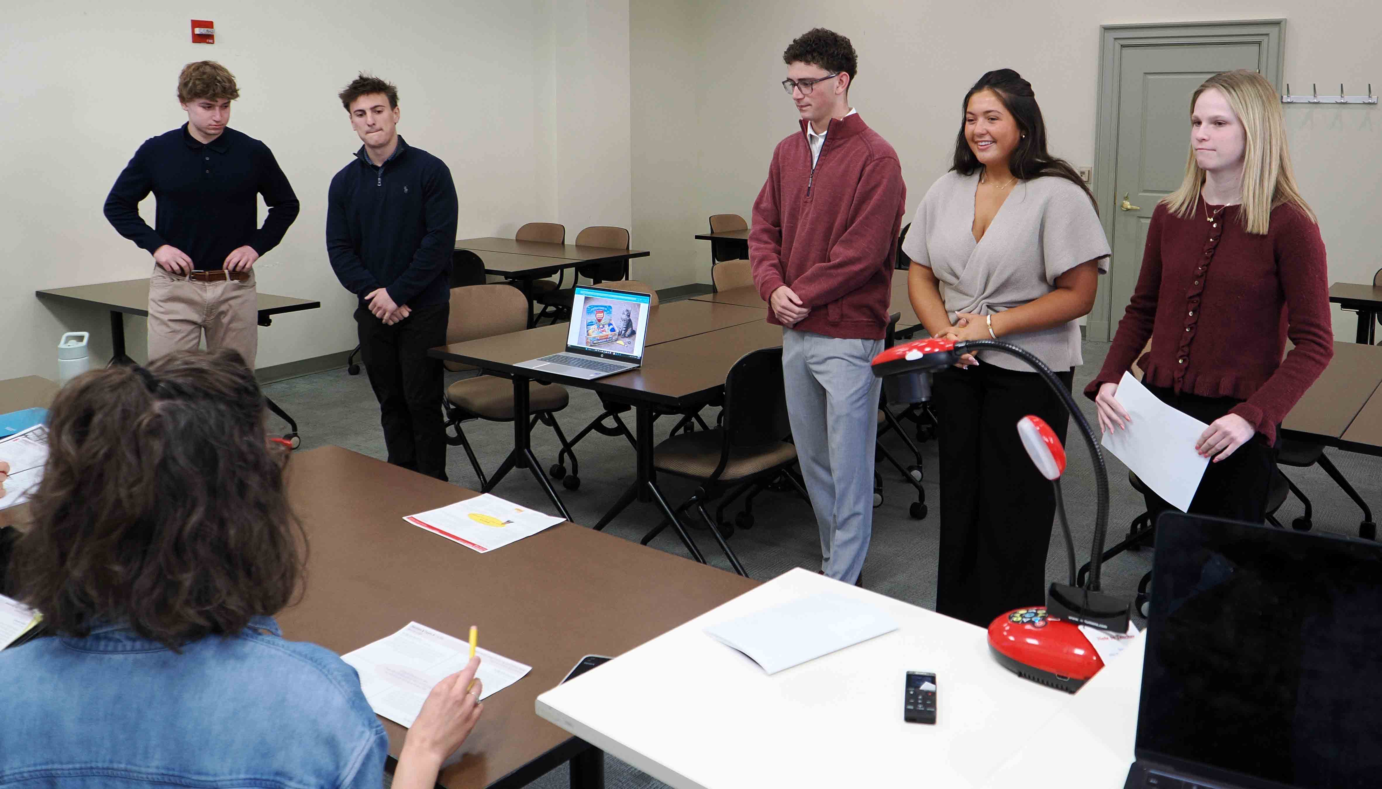 Four students stand at the front of a classroom giving a presentation, with a laptop open on a table and an audience member taking notes in the foreground.