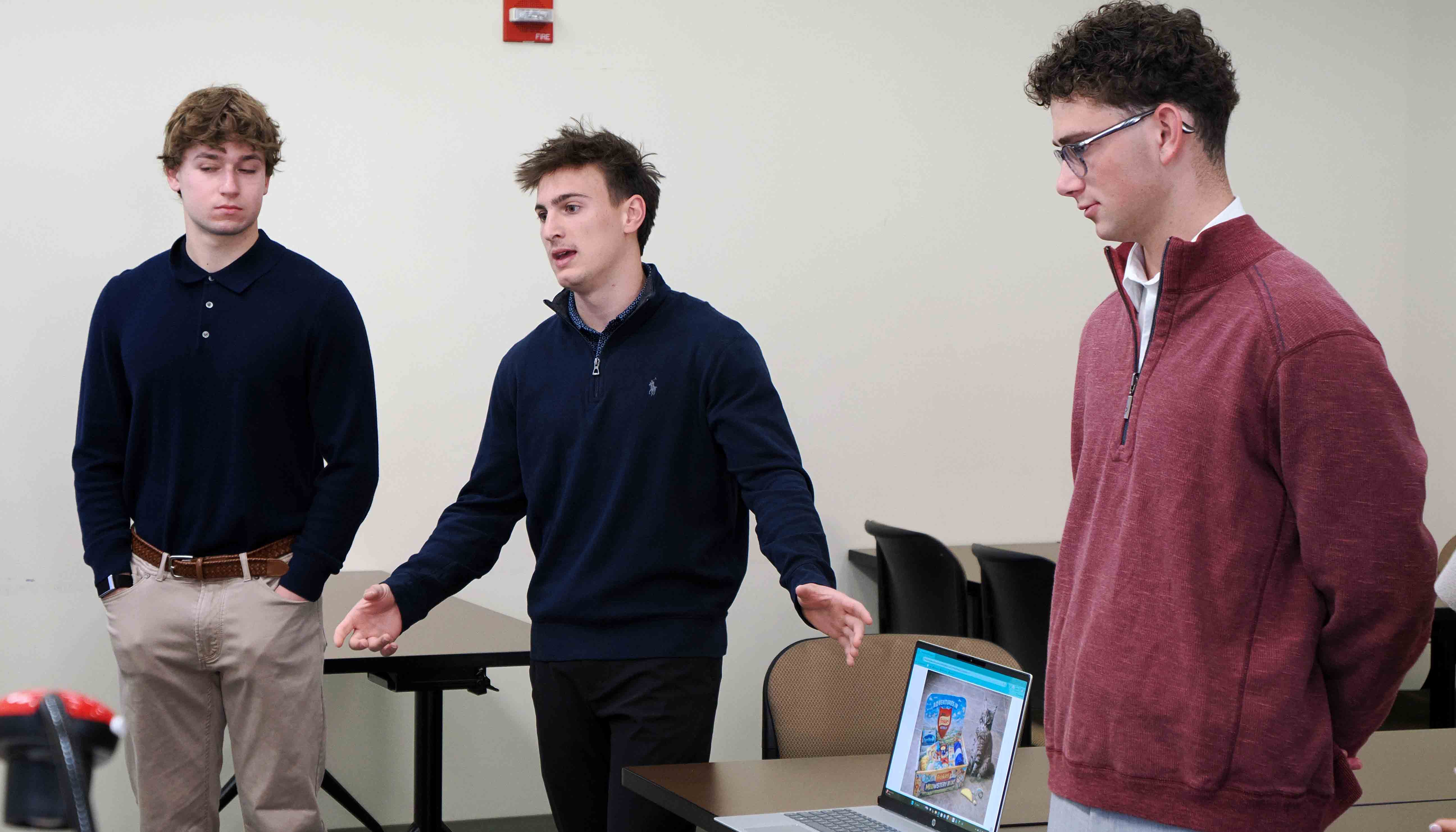 Three students stand at the front of a classroom, with one gesturing while speaking beside an open laptop showing a presentation slide.
