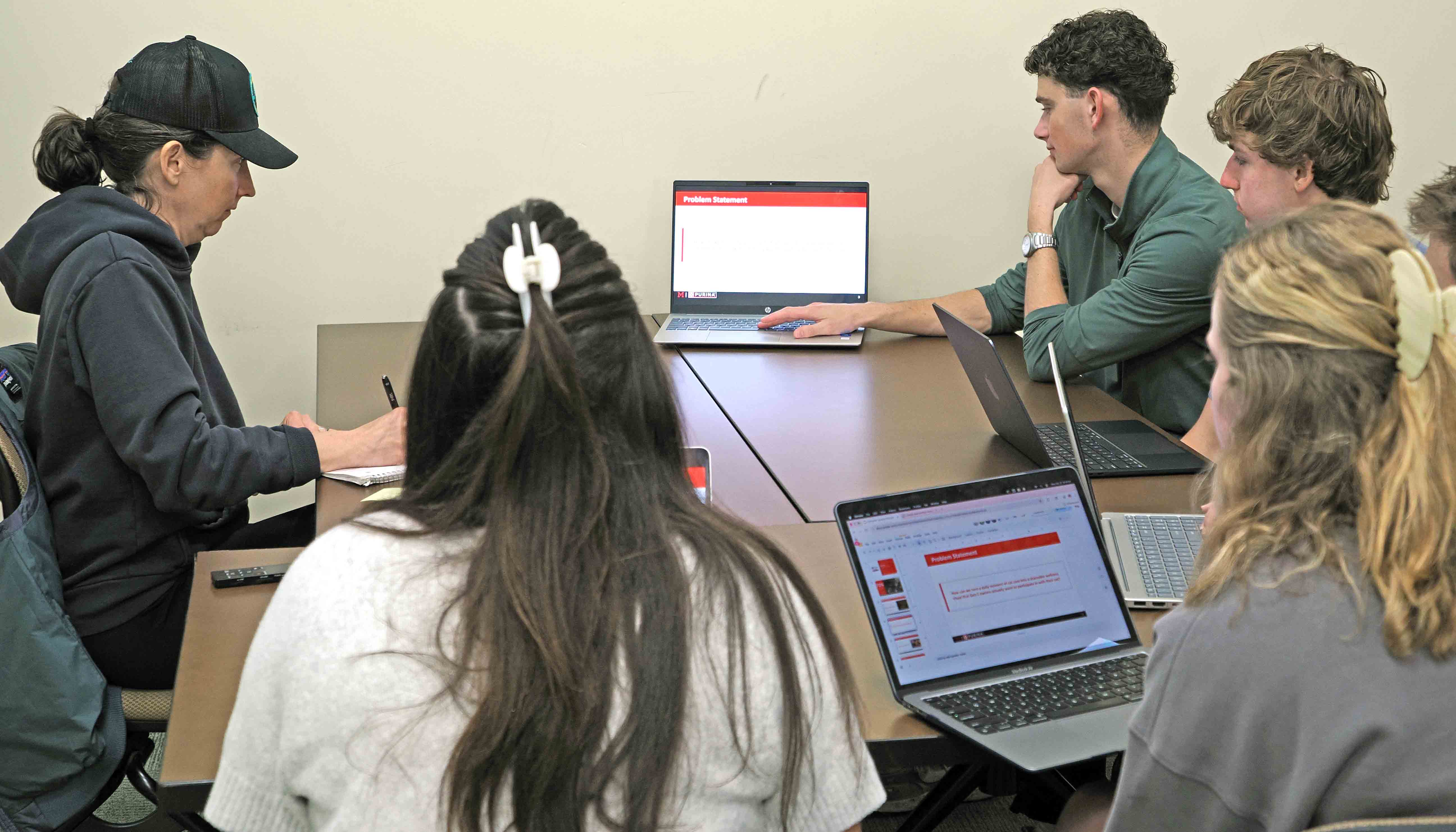 Small group meeting around a table, with students using laptops while one person presents a "Problem Statement" slide on a laptop screen.