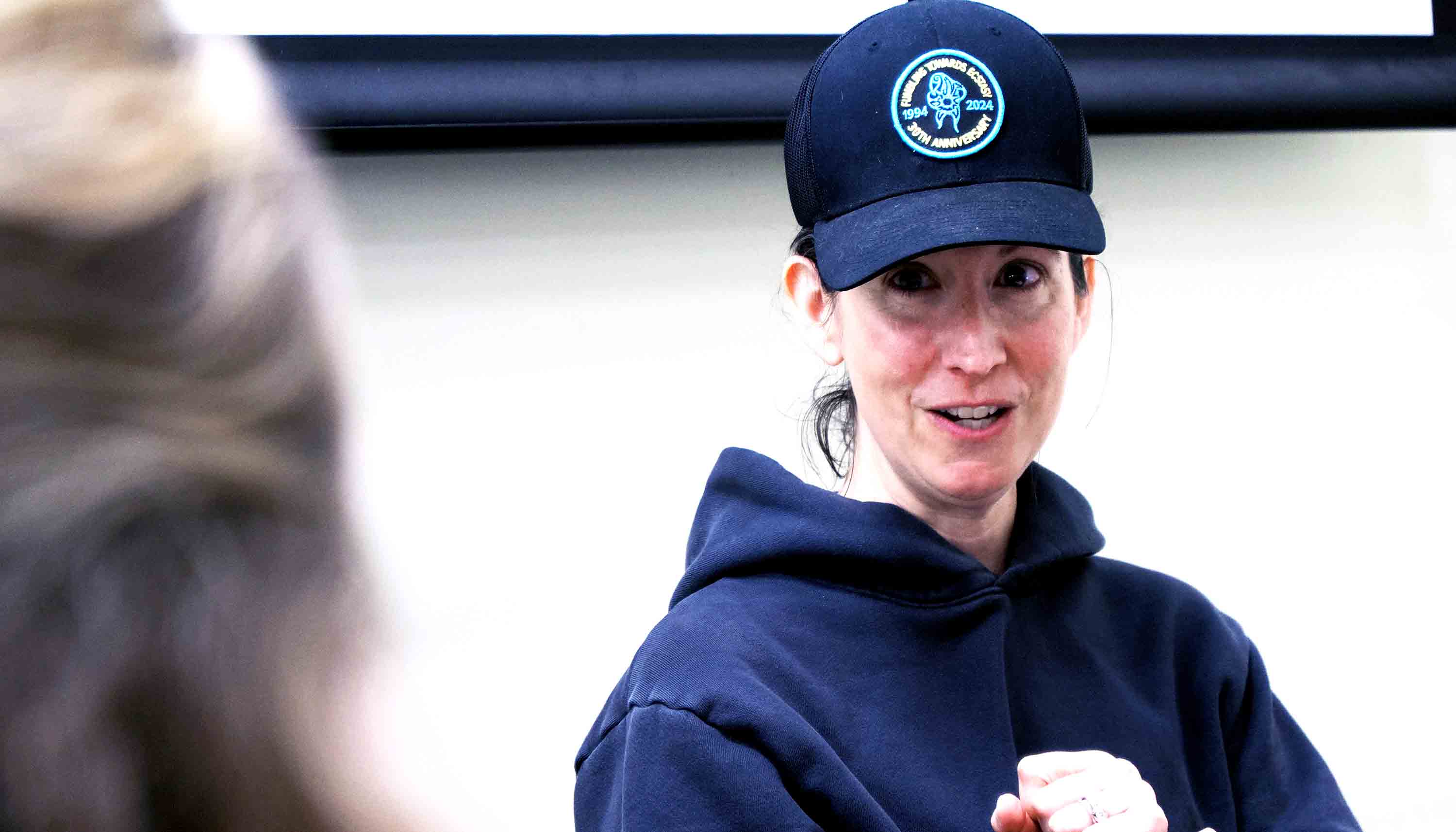Woman in a navy hoodie and black baseball cap with a circular anniversary patch, speaking while holding her hands up in a boxing stance.