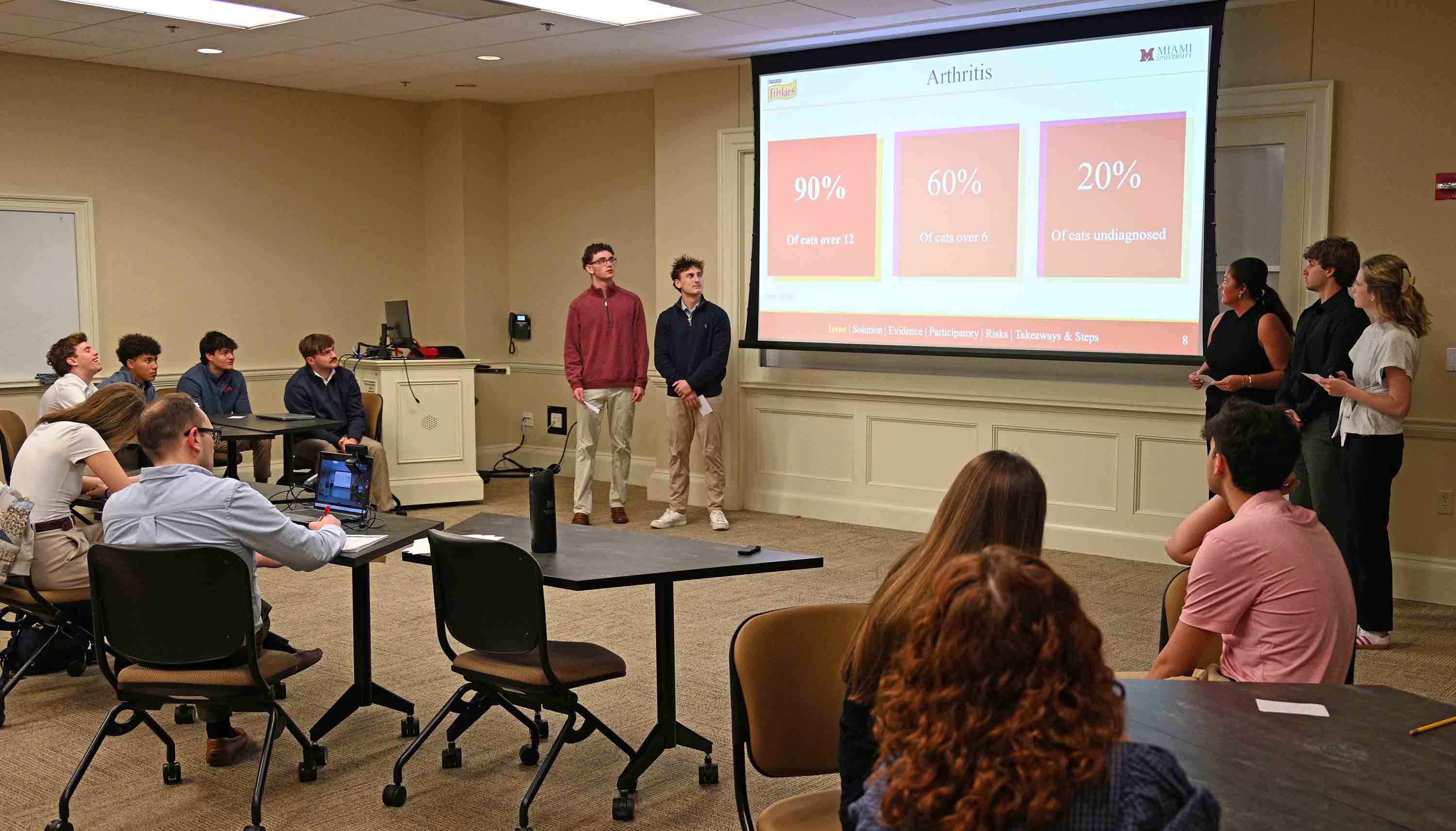 Students give a classroom presentation while a slide titled "Arthritis" with percentage statistics is projected on a screen as classmates watch.