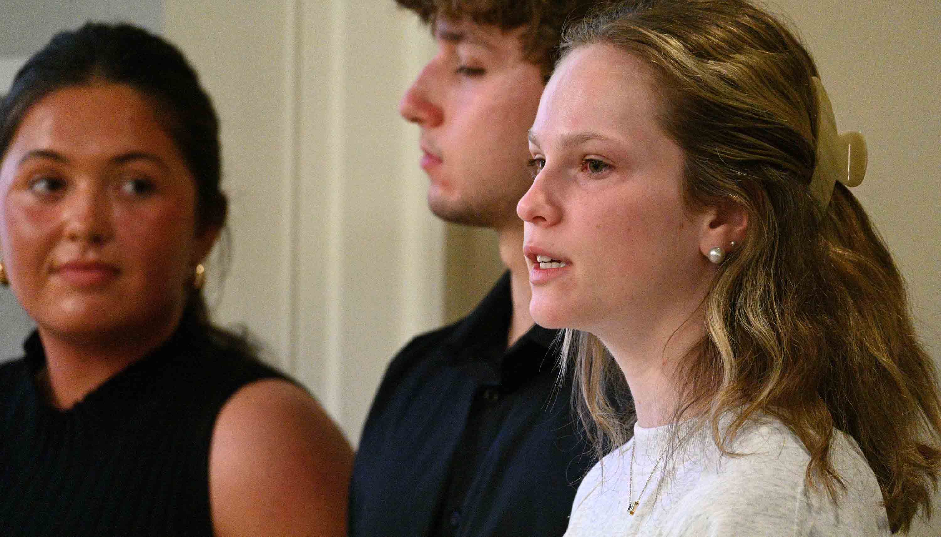 Close-up of a young woman speaking while two other people sit slightly out of focus behind her in an indoor meeting setting.