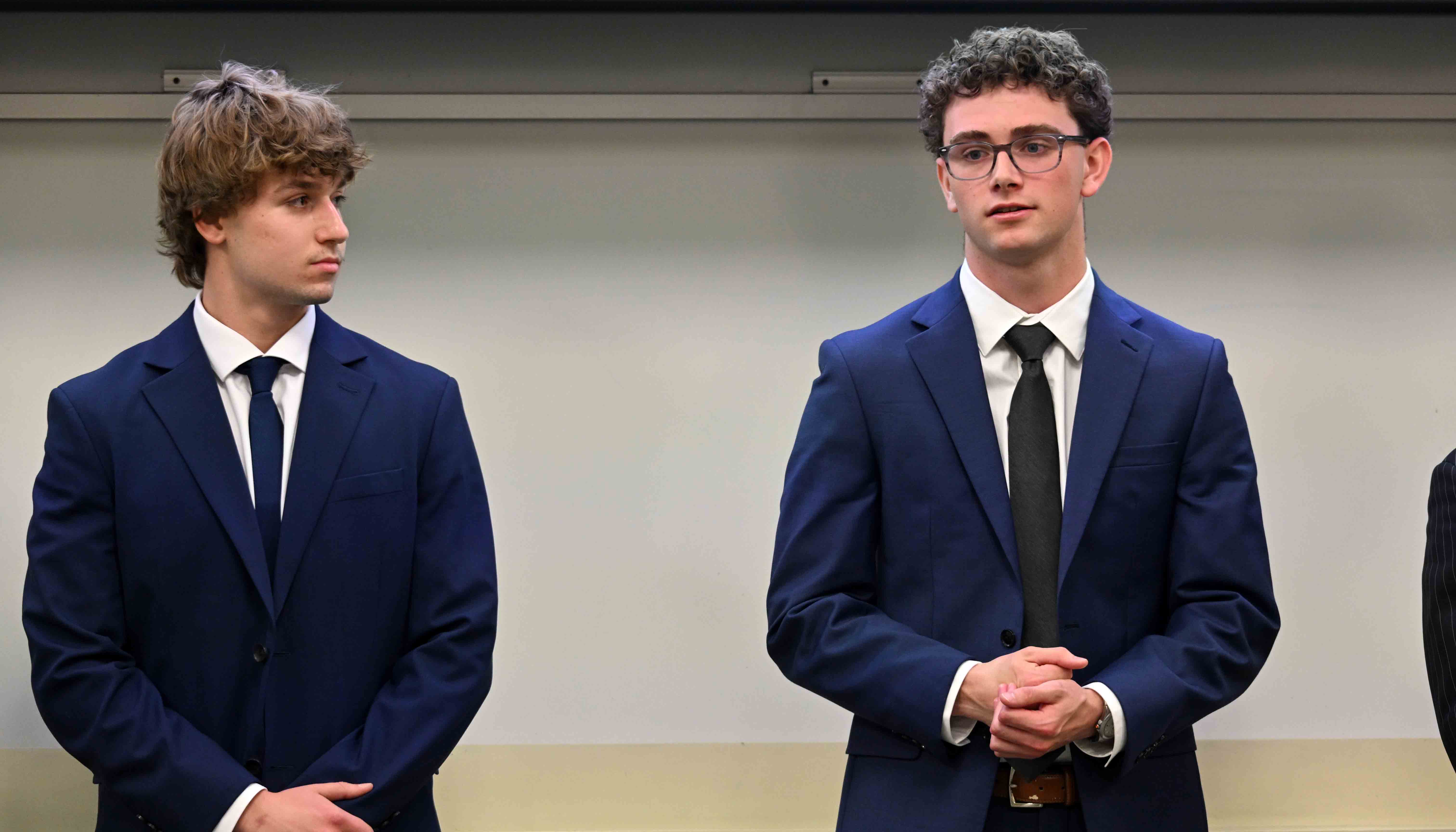 Two young men in blue suits and ties stand indoors against a plain wall, one wearing glasses and clasping his hands while the other looks toward him.
