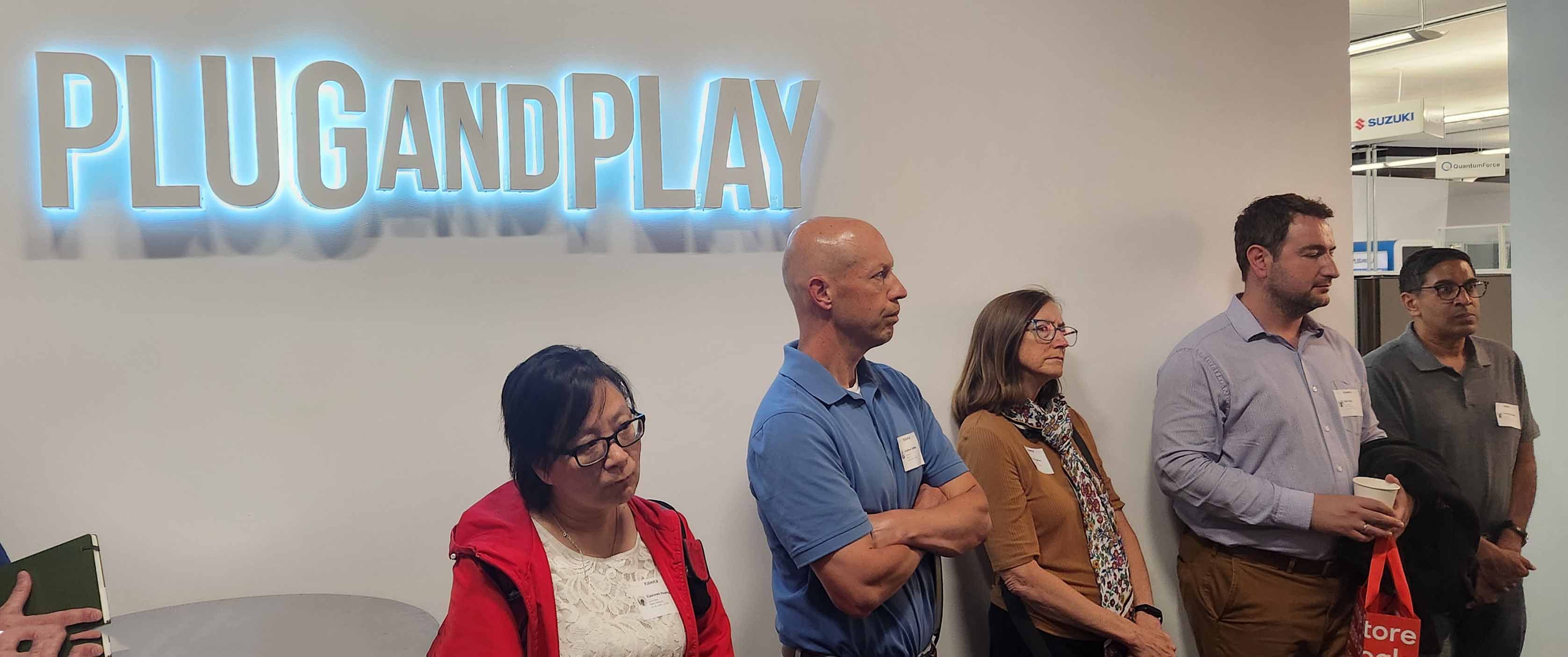 Attendees stand listening beneath a glowing “PLUG AND PLAY” sign at an indoor event.