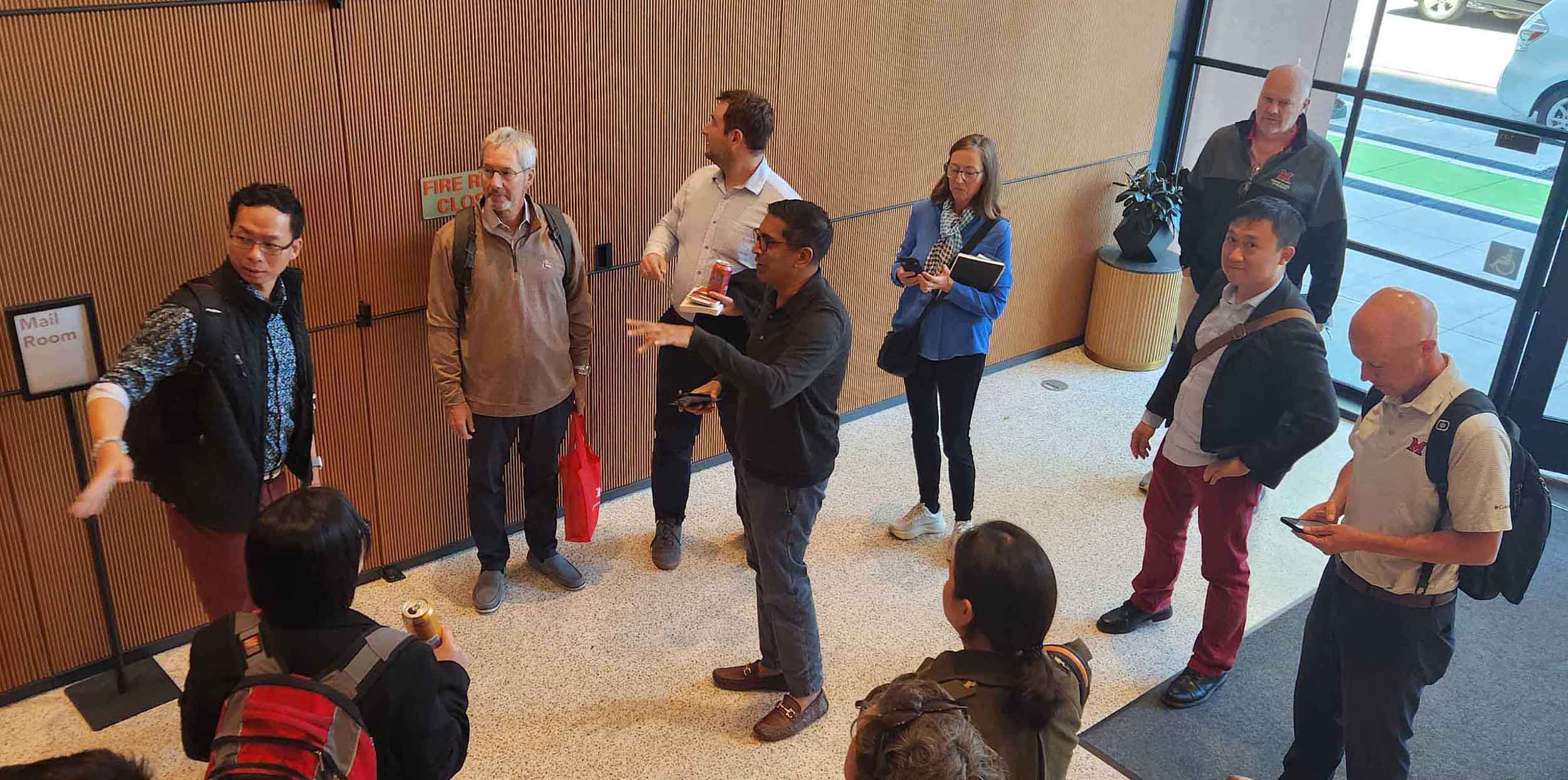 Group of adults gathered in a building lobby, listening as one person gestures and speaks near the entrance doors.