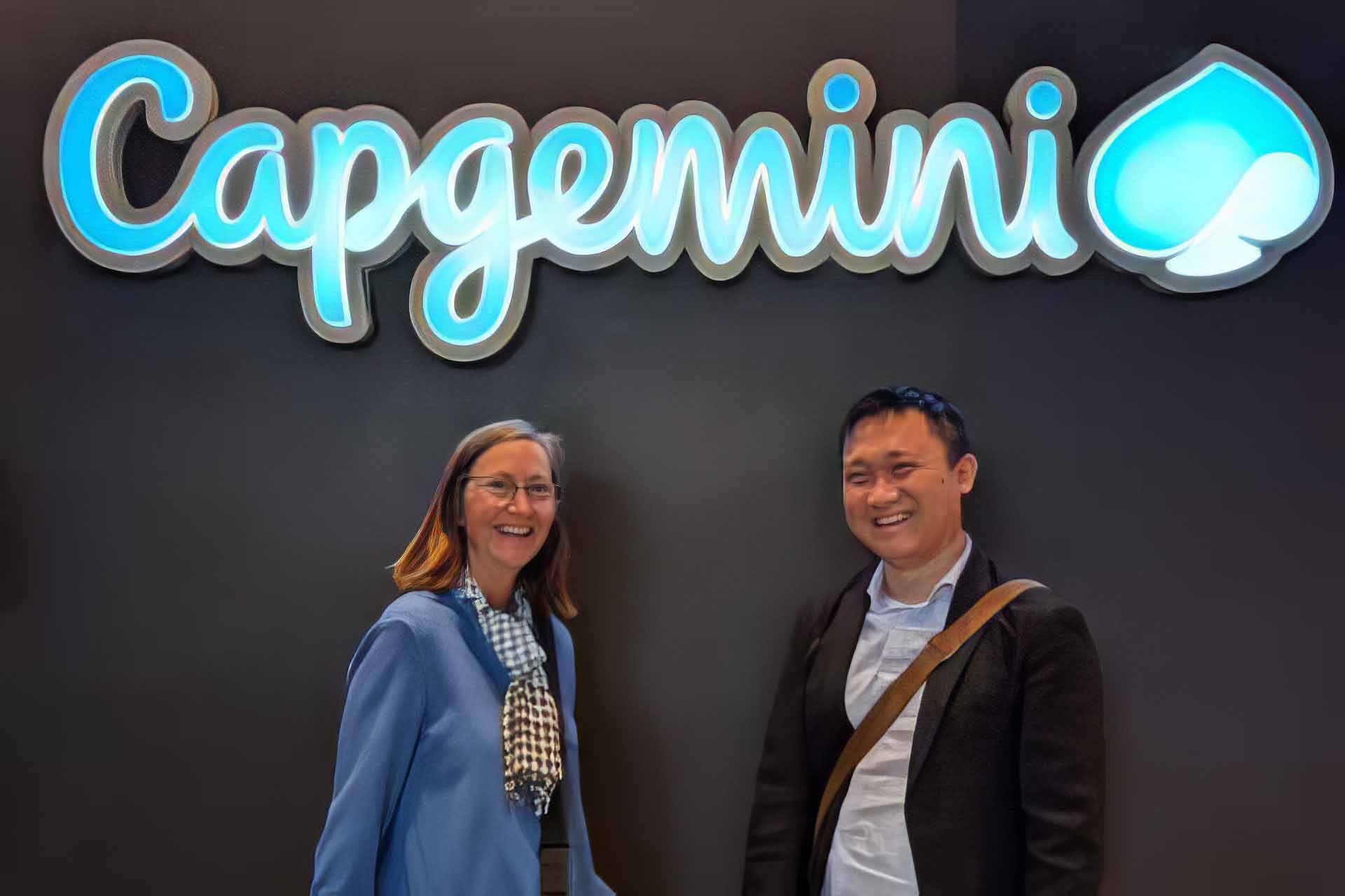 Two people smiling in front of a wall with a large illuminated "Capgemini" sign and logo above them.