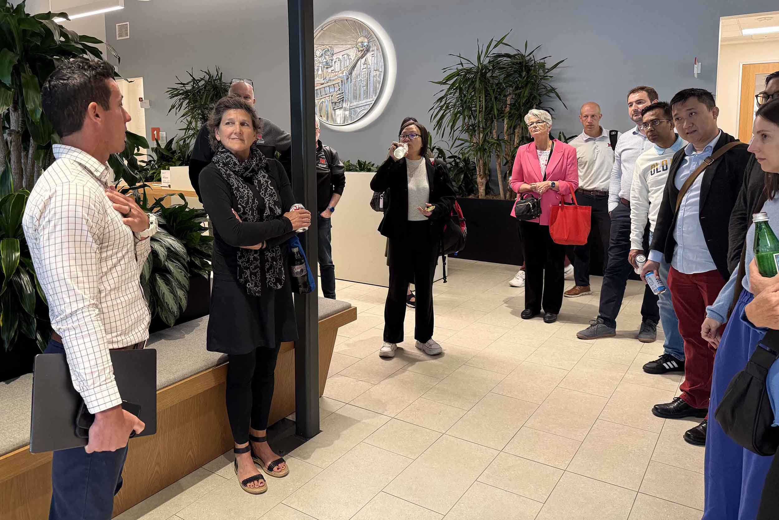 A group of people stand in an office lobby, listening to a man speaking at left, with indoor plants and a large wall medallion in the background.