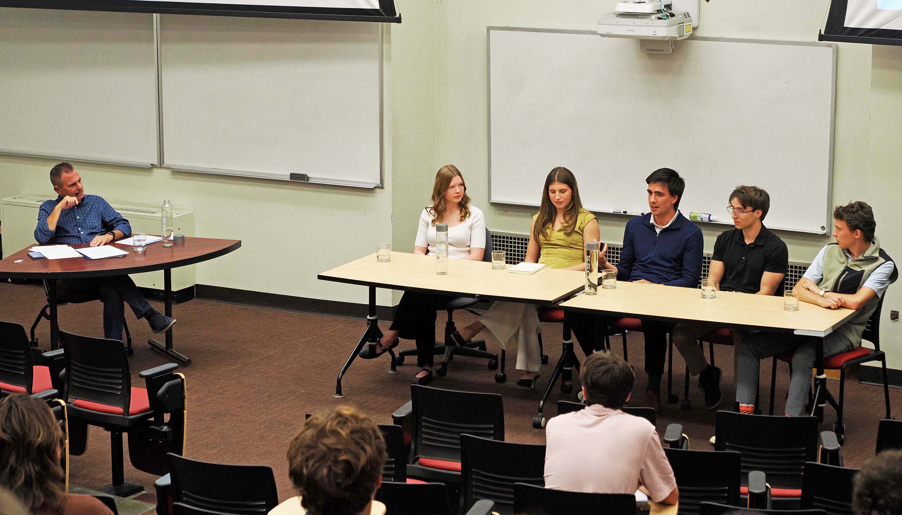 Panel discussion in a classroom, with a moderator at a separate table and five speakers seated behind desks facing an audience.