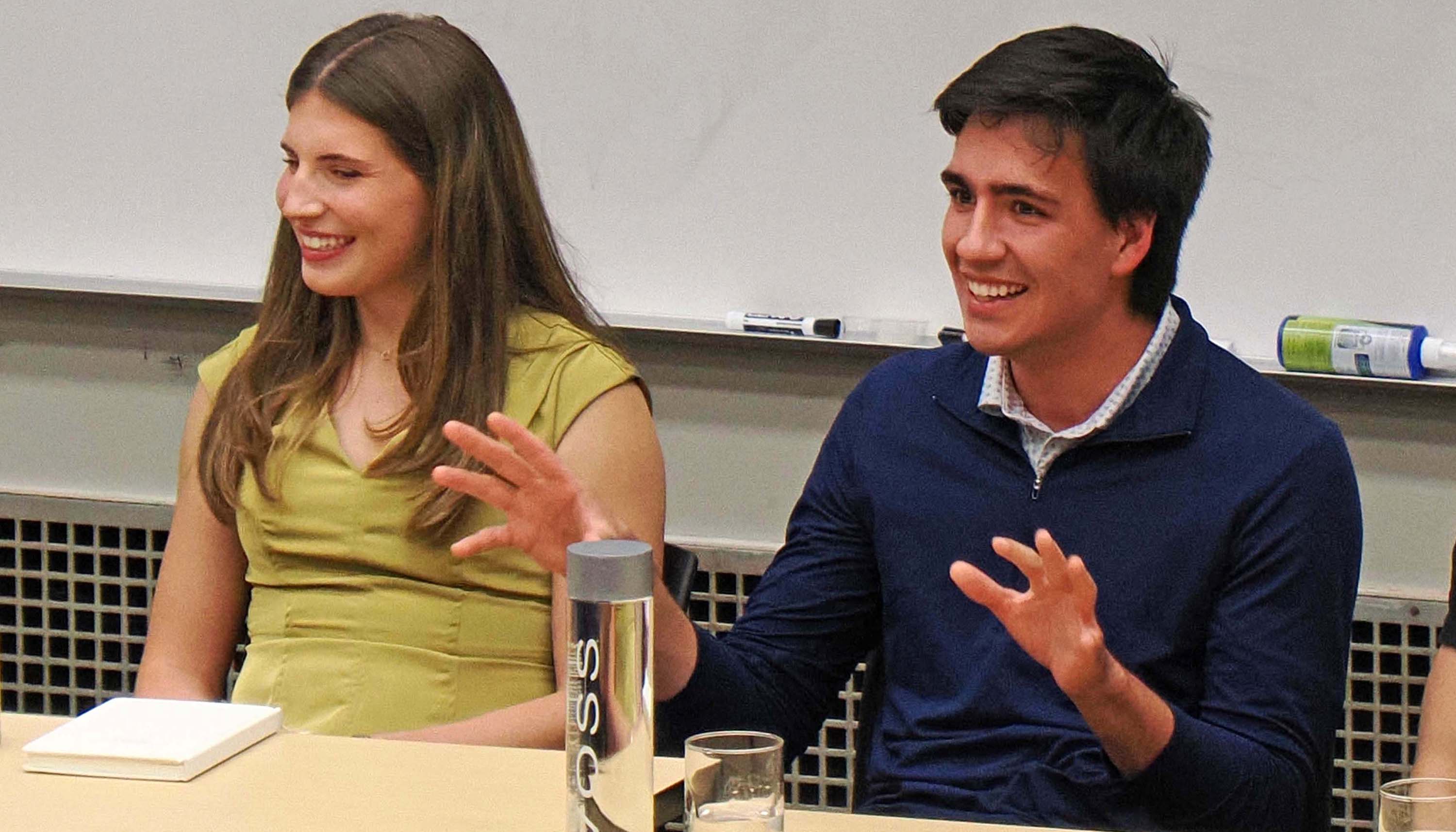 Two people sit at a table in a classroom, smiling while one gestures during a discussion in front of a whiteboard.