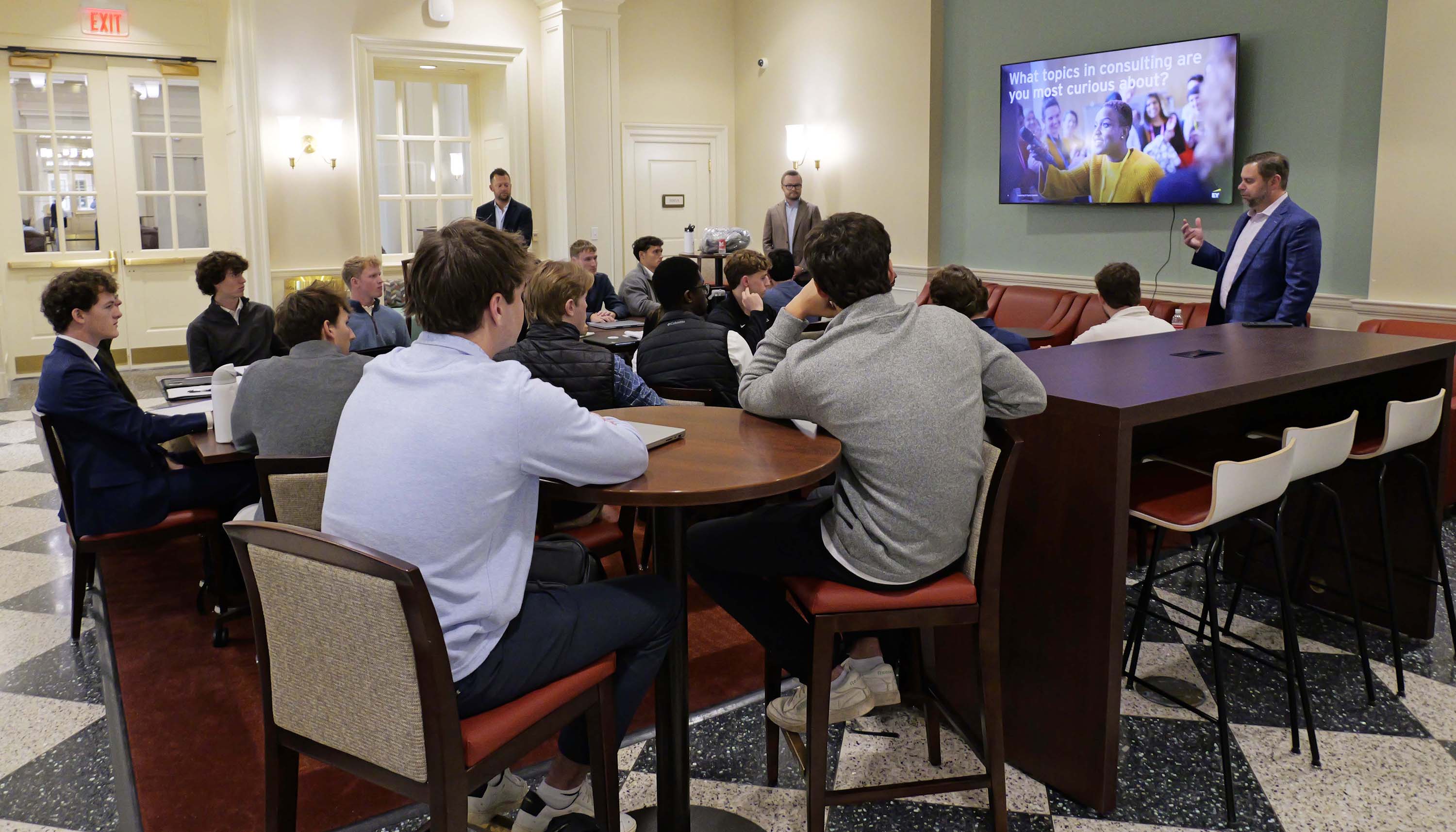 Students seated around tables listen to a speaker giving a consulting presentation, with a slide displayed on a wall-mounted screen.