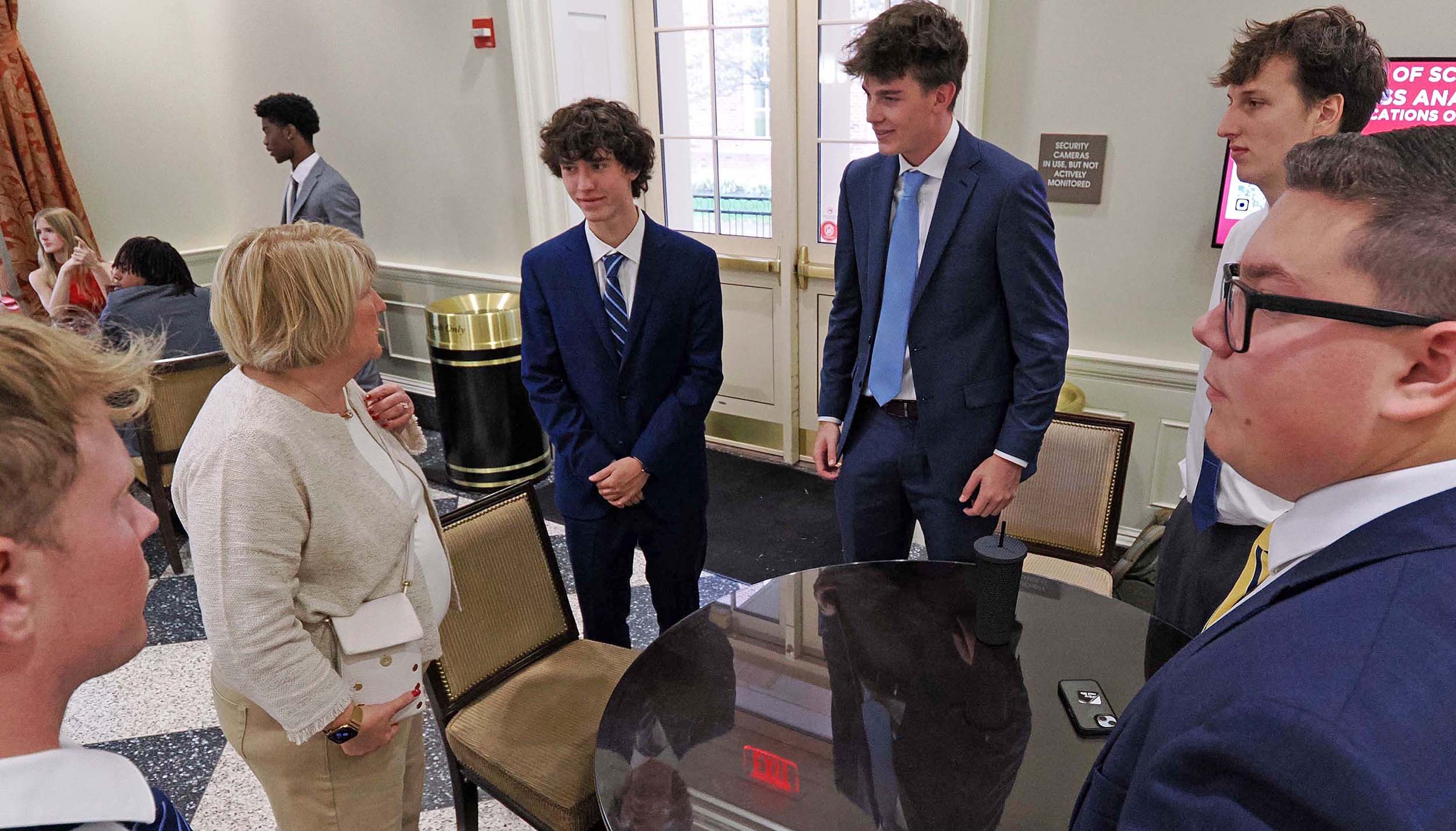 Group of young men in suits chatting with an older woman around a small table in an indoor lobby or reception area.