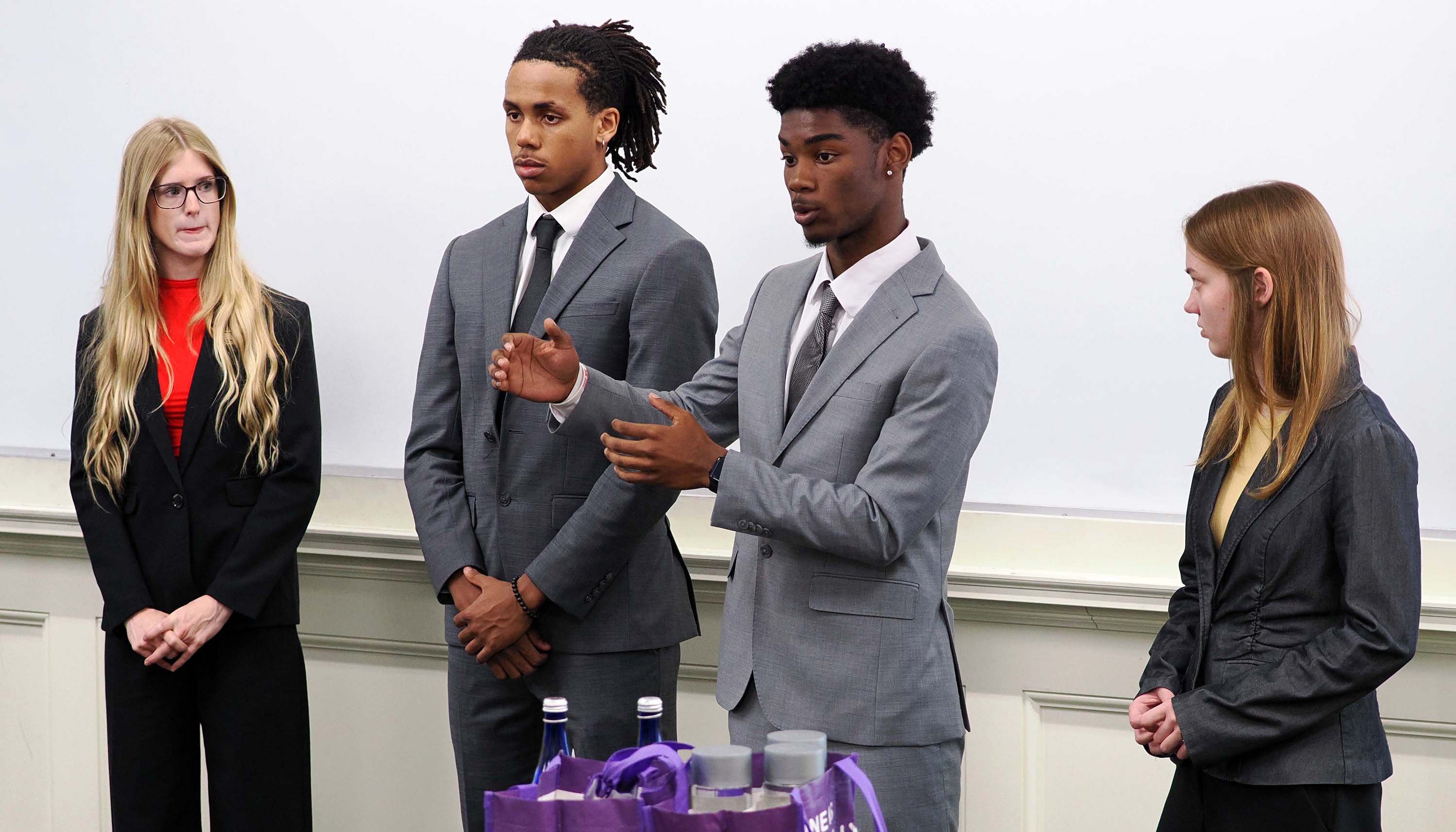 Four young adults in business attire stand in front of a whiteboard, with one person gesturing as if giving a presentation.