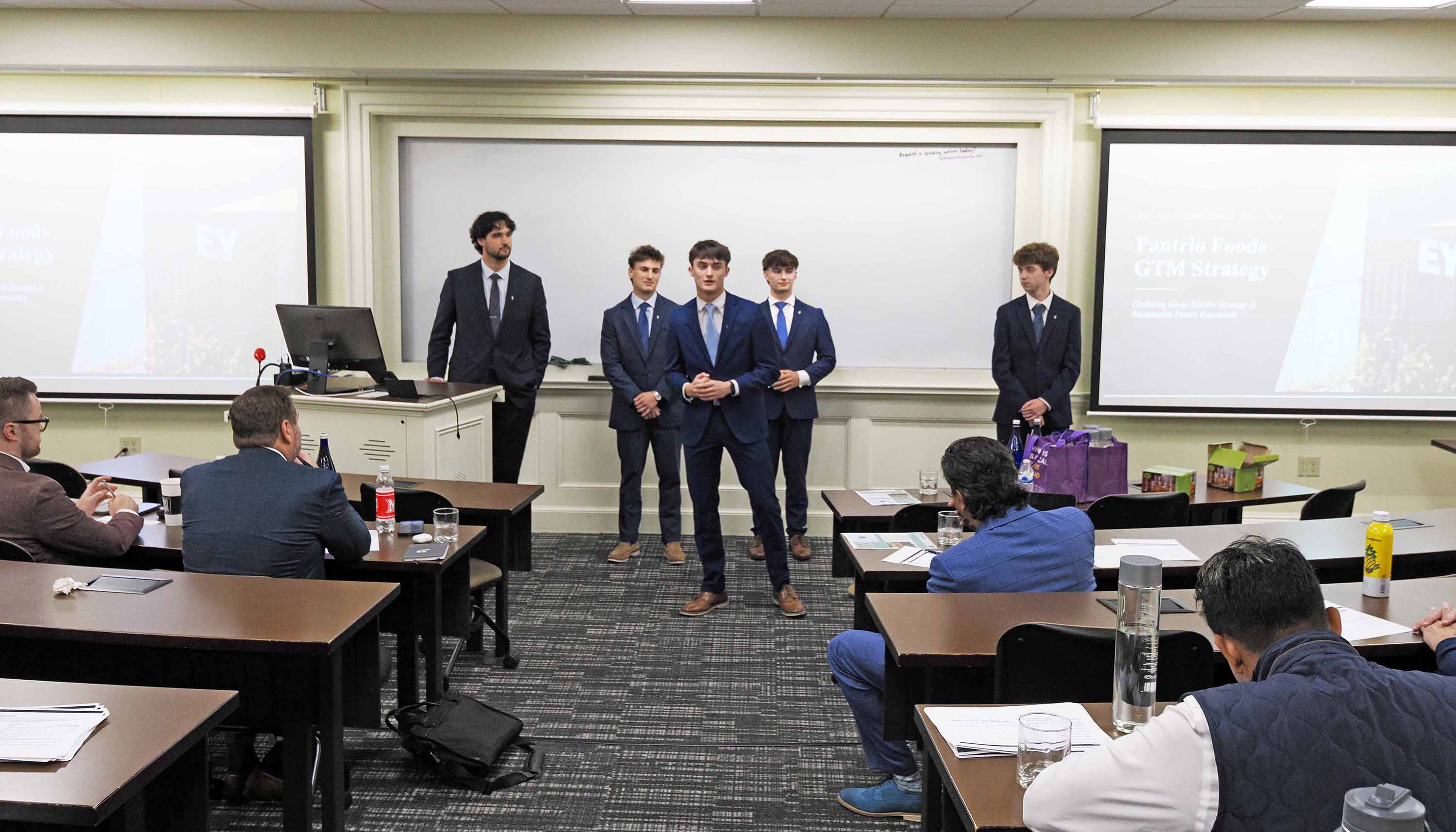Five presenters in suits stand at the front of a classroom delivering a business presentation to seated attendees, with slides projected on screens.