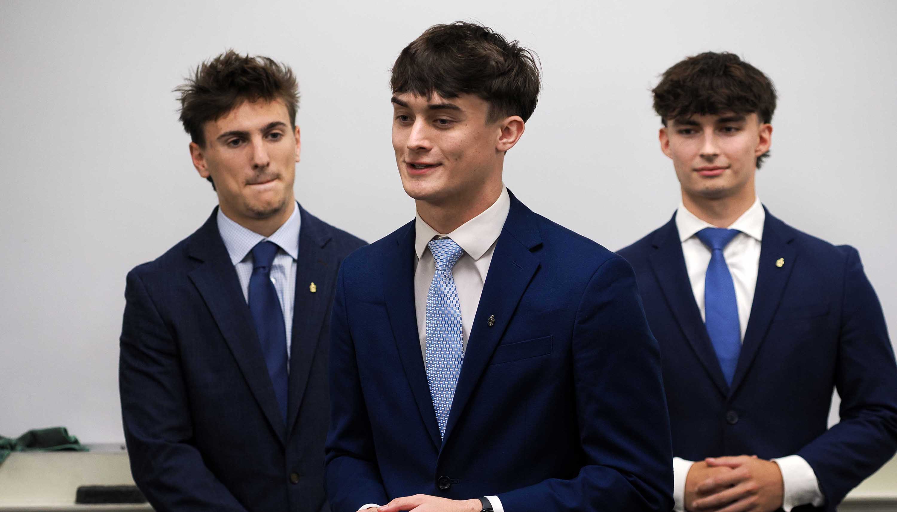 Three young men in navy suits and ties stand in a row, with the one in front speaking while the others look on.