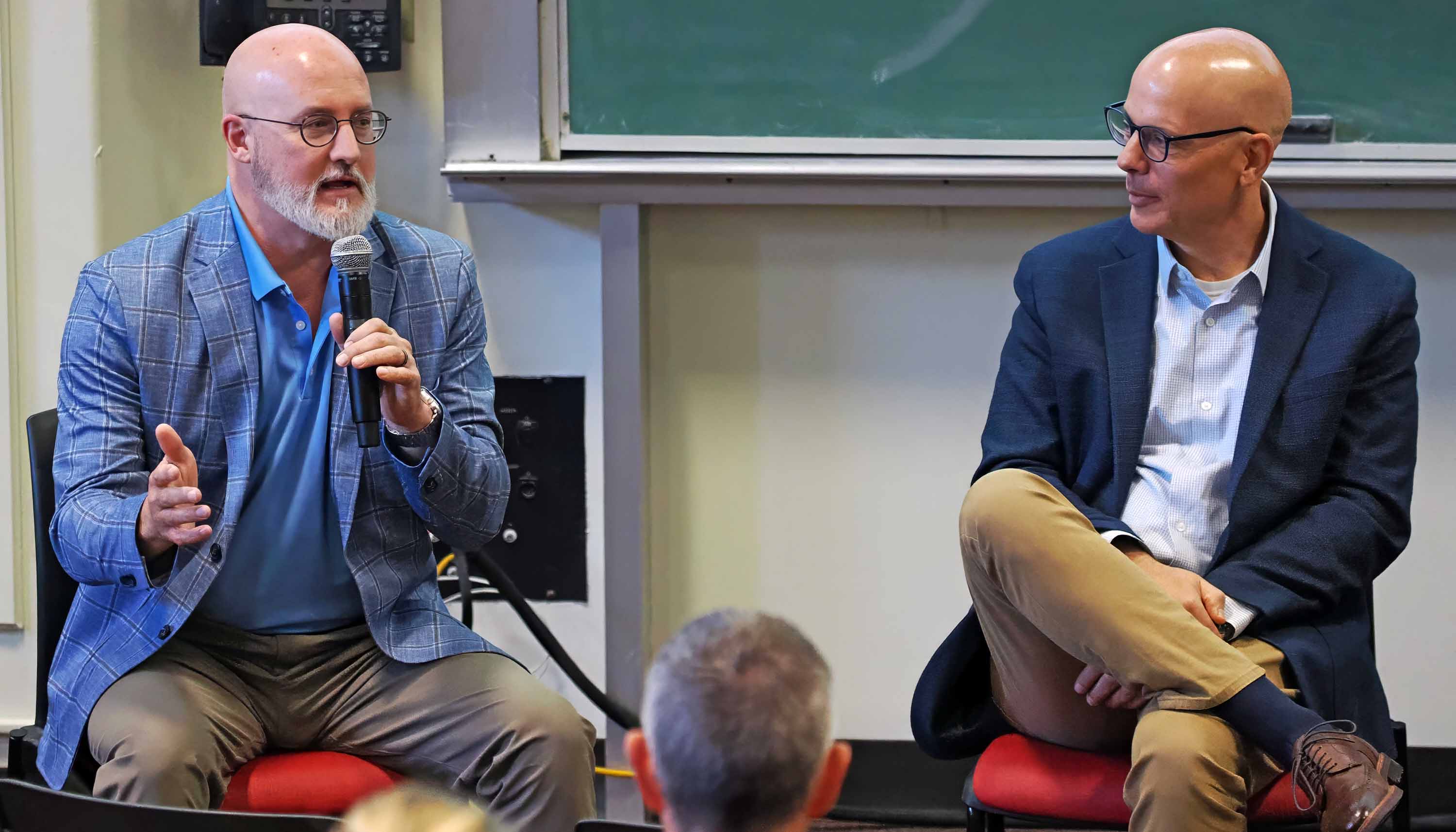 Two men in business attire sit on chairs during a talk; one speaks into a handheld microphone while the other listens, with a chalkboard behind them.