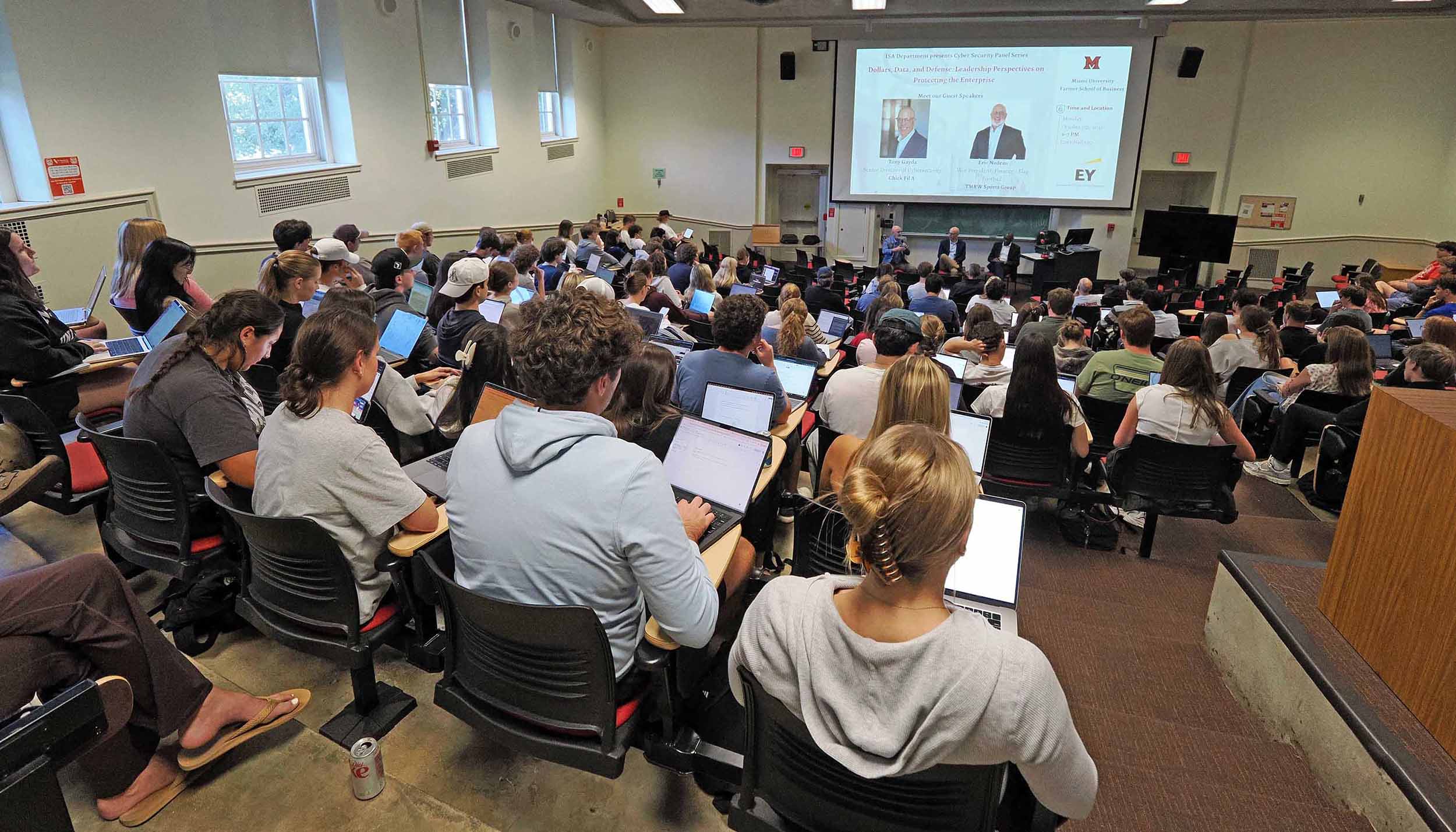 Students in a packed lecture hall using laptops while a panel discussion is presented on a large projection screen at the front of the room.
