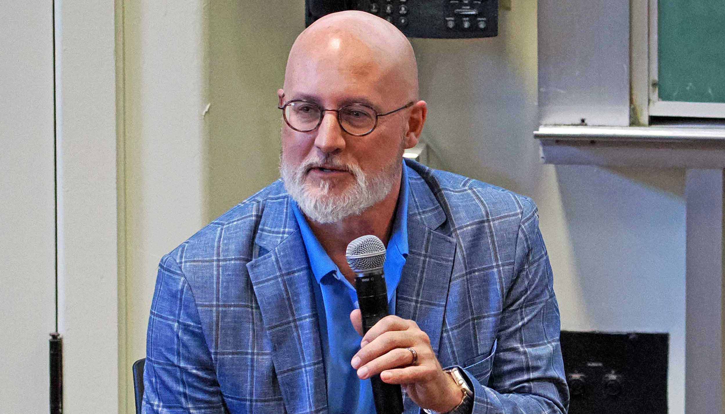 Eric Nadeau with glasses and a white beard speaks into a handheld microphone during an indoor talk or panel discussion.