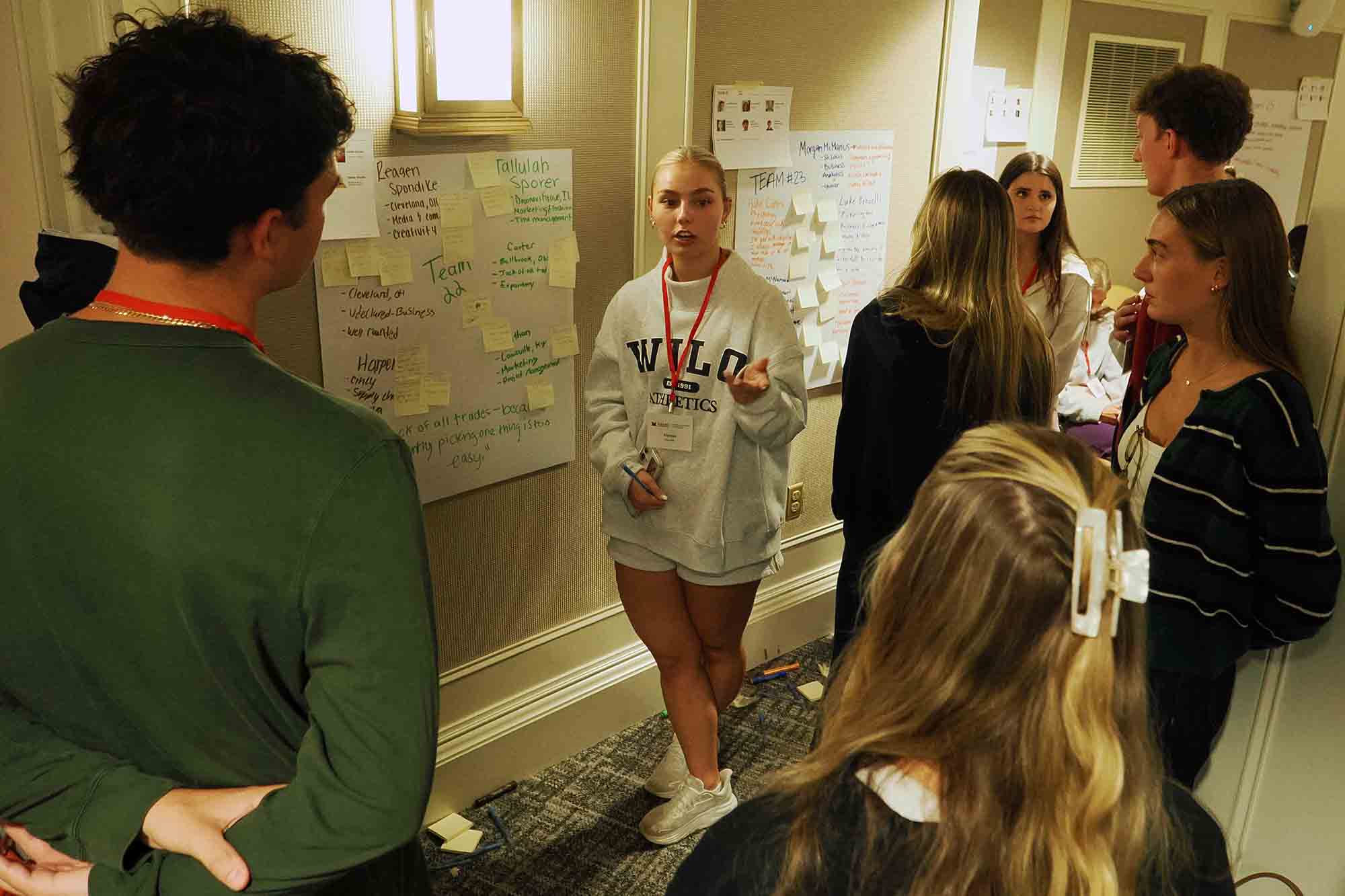 A group of students in a hallway discusses ideas while one student presents next to wall posters covered with notes and sticky notes.