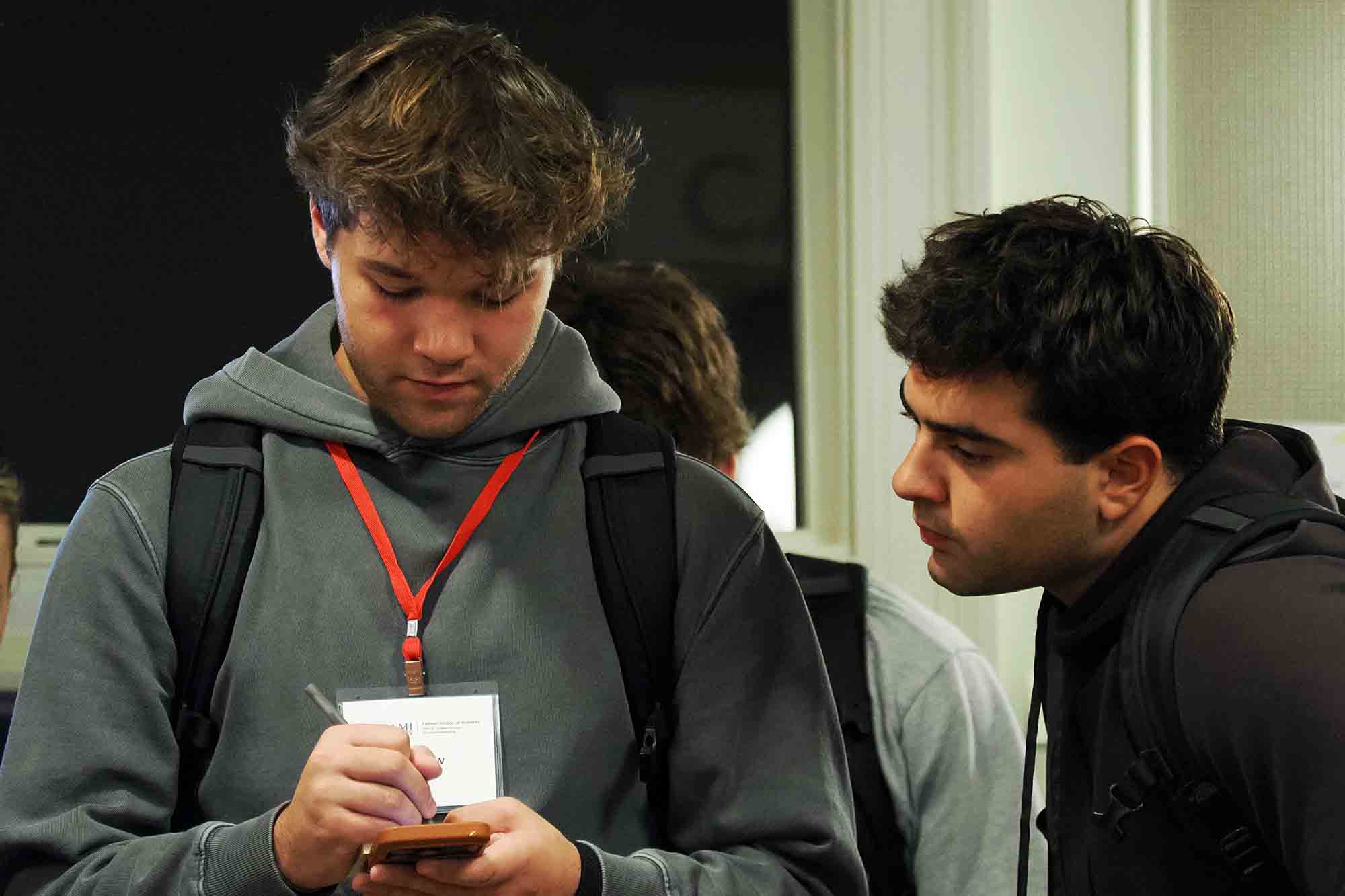 Two people wearing backpacks and conference badges; one looks down at a smartphone while the other leans in to watch.