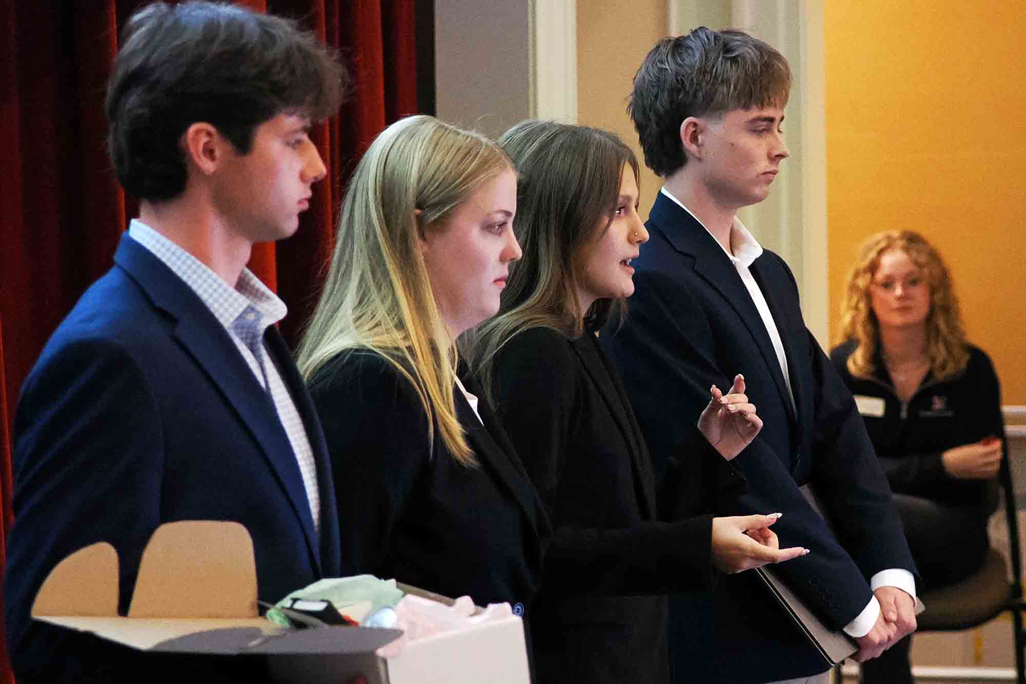Three young adults in business attire stand side by side giving a presentation indoors, with one woman gesturing while speaking.