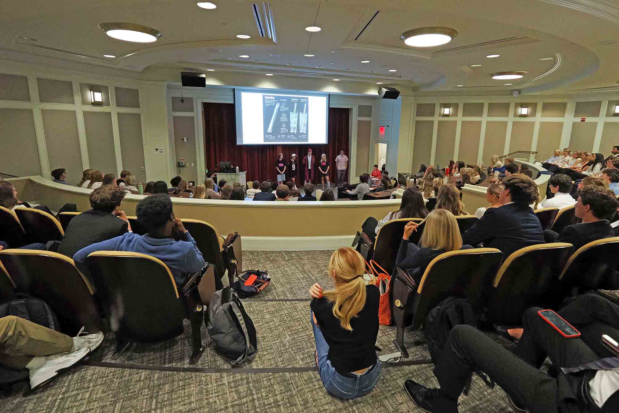 Students seated in a large lecture hall watch a group presenting at the front beneath a projected slide.