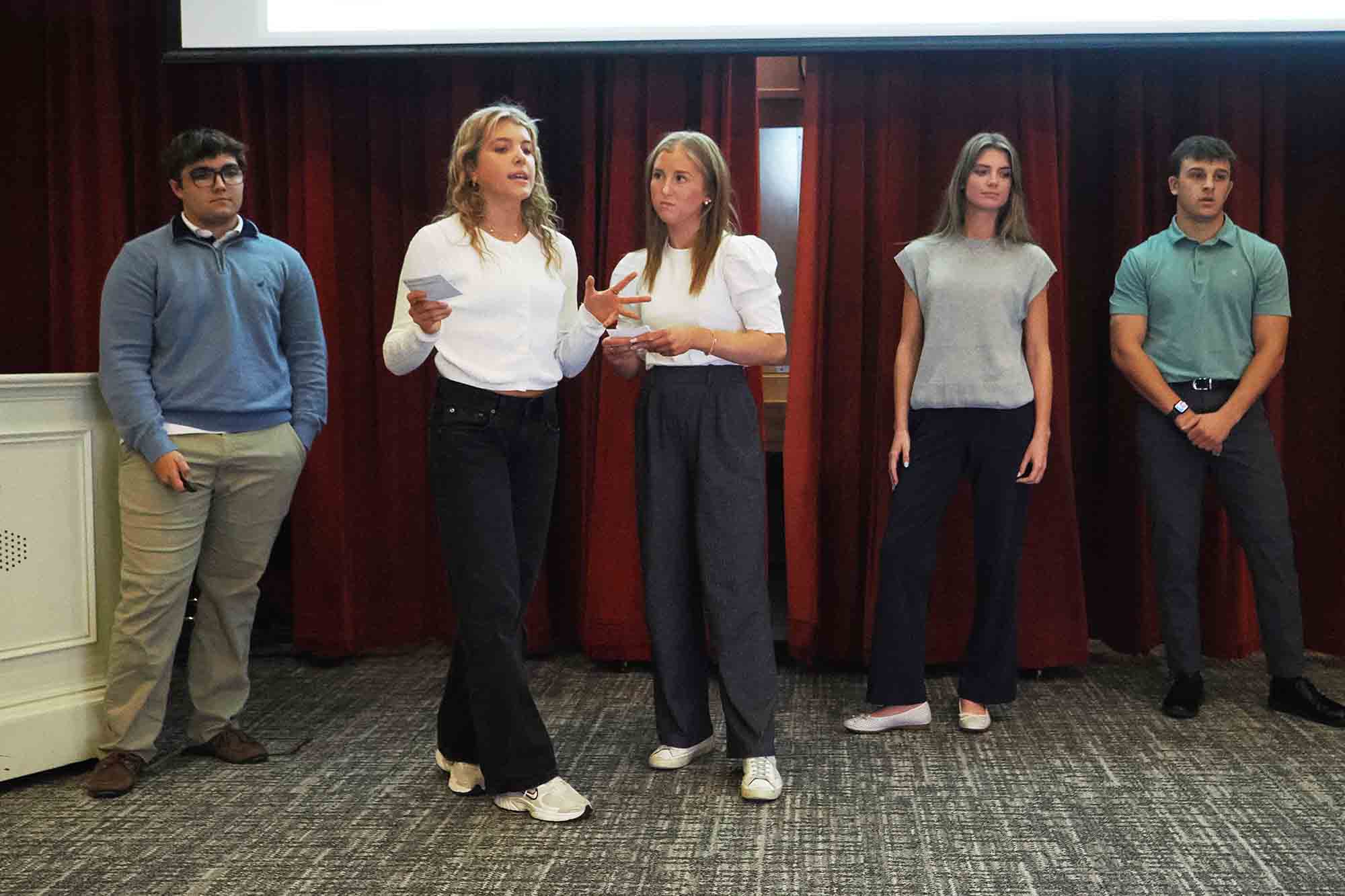 Five students stand in front of red curtains during a presentation, with two in the center holding notes and speaking.