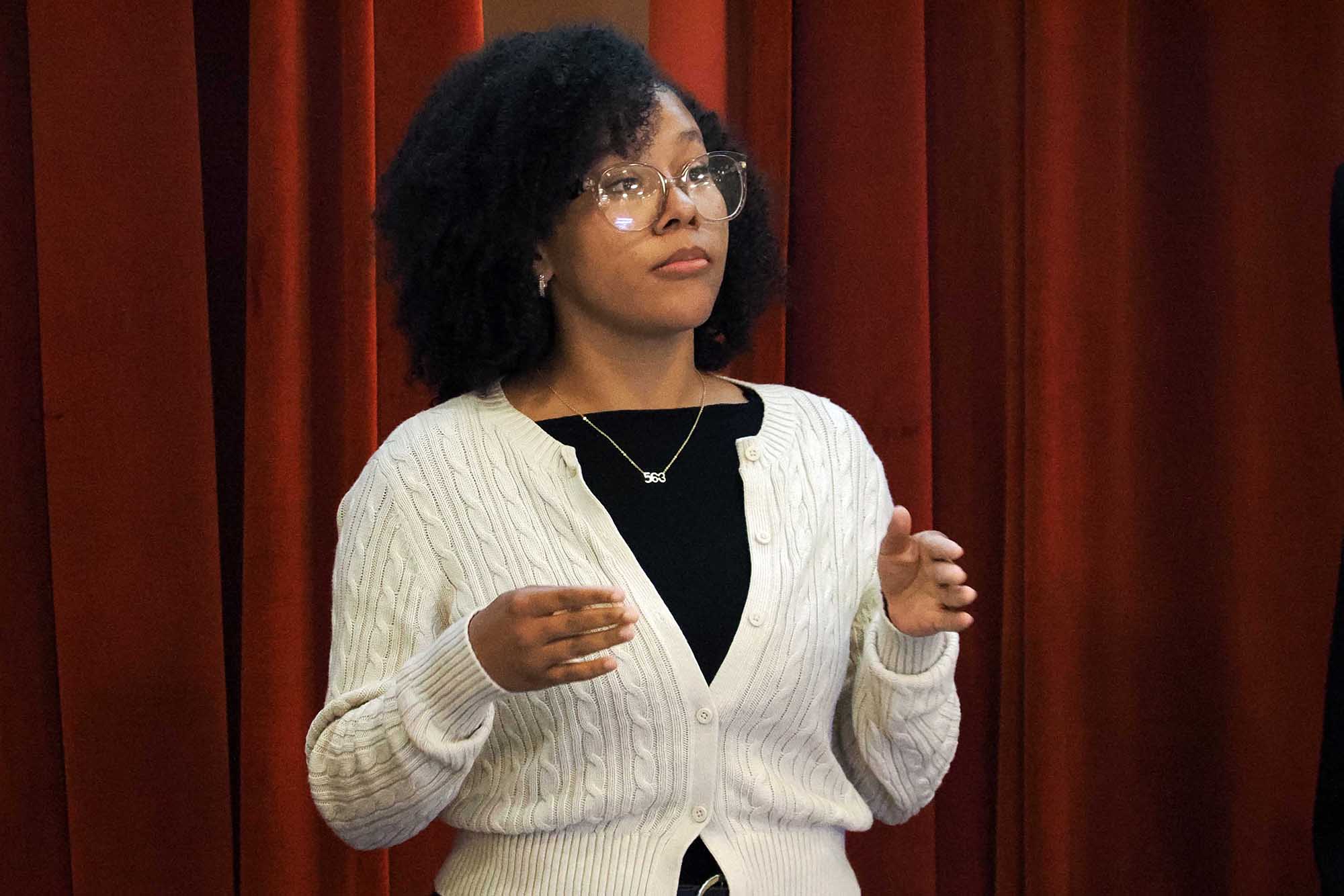 Person with curly hair and glasses gestures while speaking in front of red theater curtains.