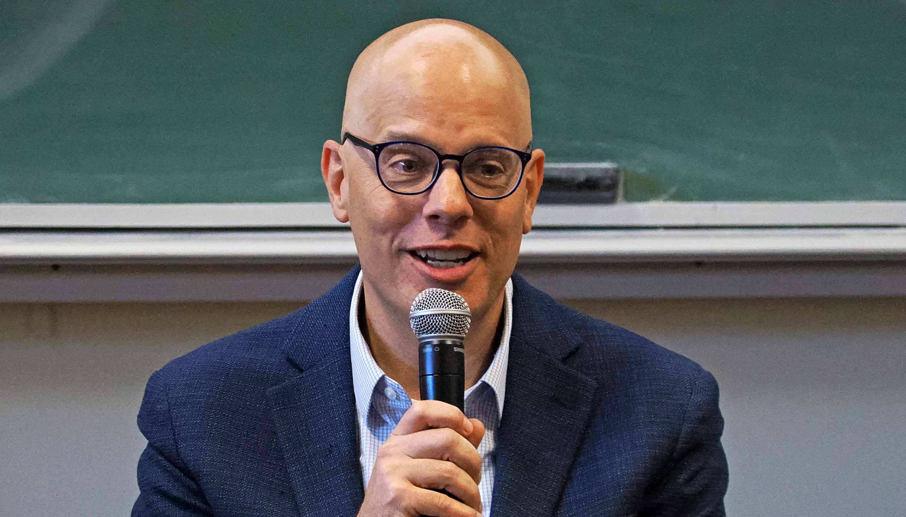 Tony Gayda, a bald man wearing glasses speaks into a handheld microphone during an indoor talk, with a chalkboard behind him.