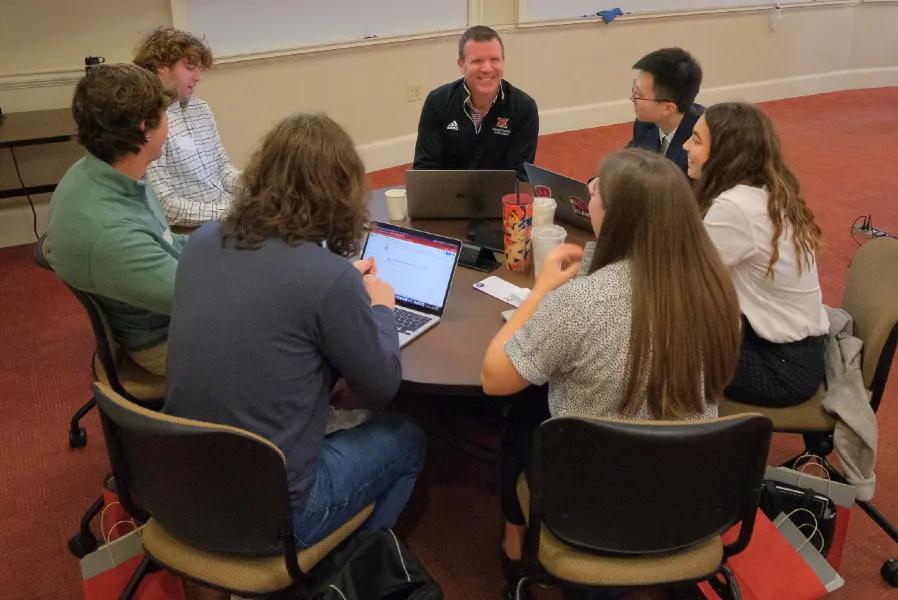 A group of students engaging in a discussion around a circular table, with laptops open and a facilitator guiding the conversation.