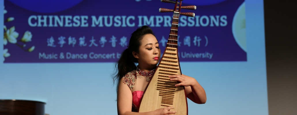 Chinese woman on stage playing an instrument with a presentation slide behind her that says "Chinese Music Impressions"