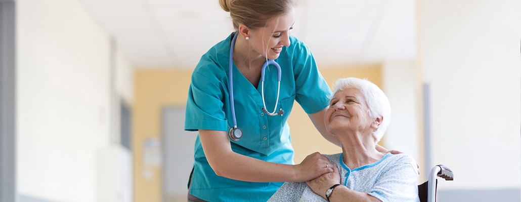 A woman in scrubs clasps hands with an elderly woman in a wheelchair
