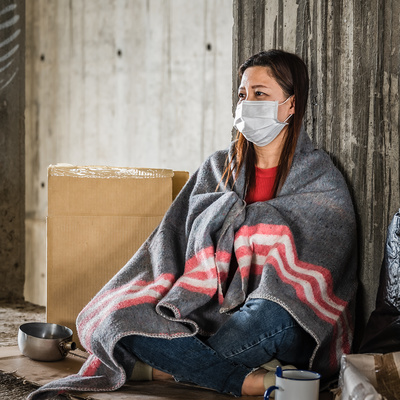 A homeless woman wearing a face mask for COVID sits under a blanket