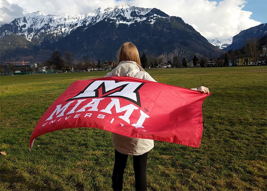 A student faces away from the camera and gazes at mountains. They hold a Miami flag behind them like a cape