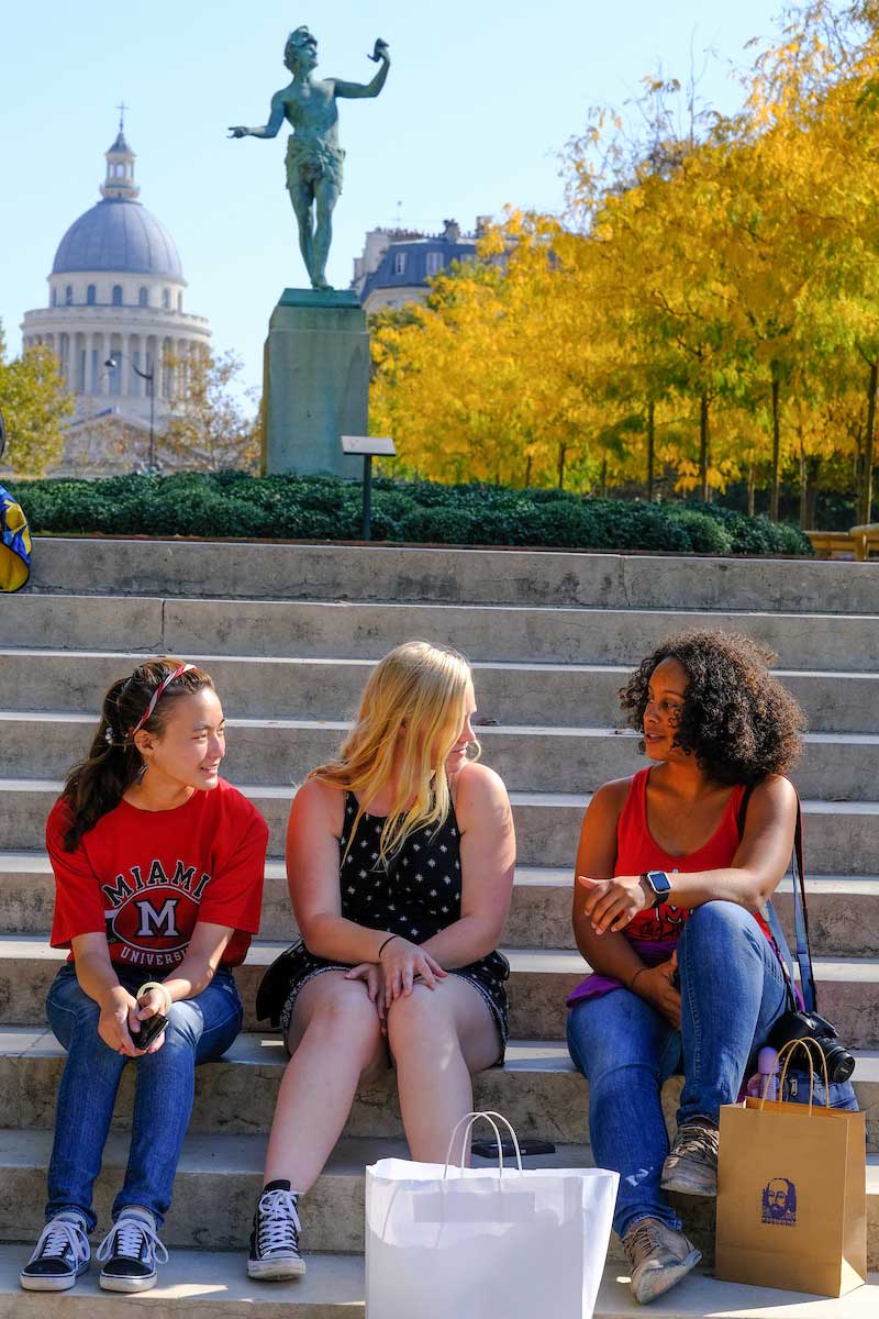 Three students sit on outdoor steps in front of a statue and dome in Paris, talking and smiling. Two wear Miami University shirts. Autumn trees with golden leaves line the background, and shopping bags sit at their feet.