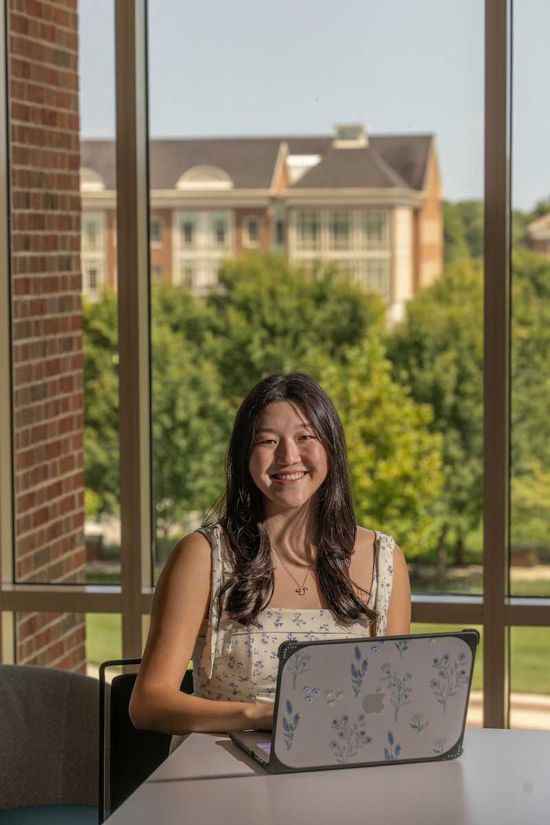 A student sits at a table using a laptop with a floral case. She is indoors near large windows with a view of trees and a brick campus building in the background on a sunny day.