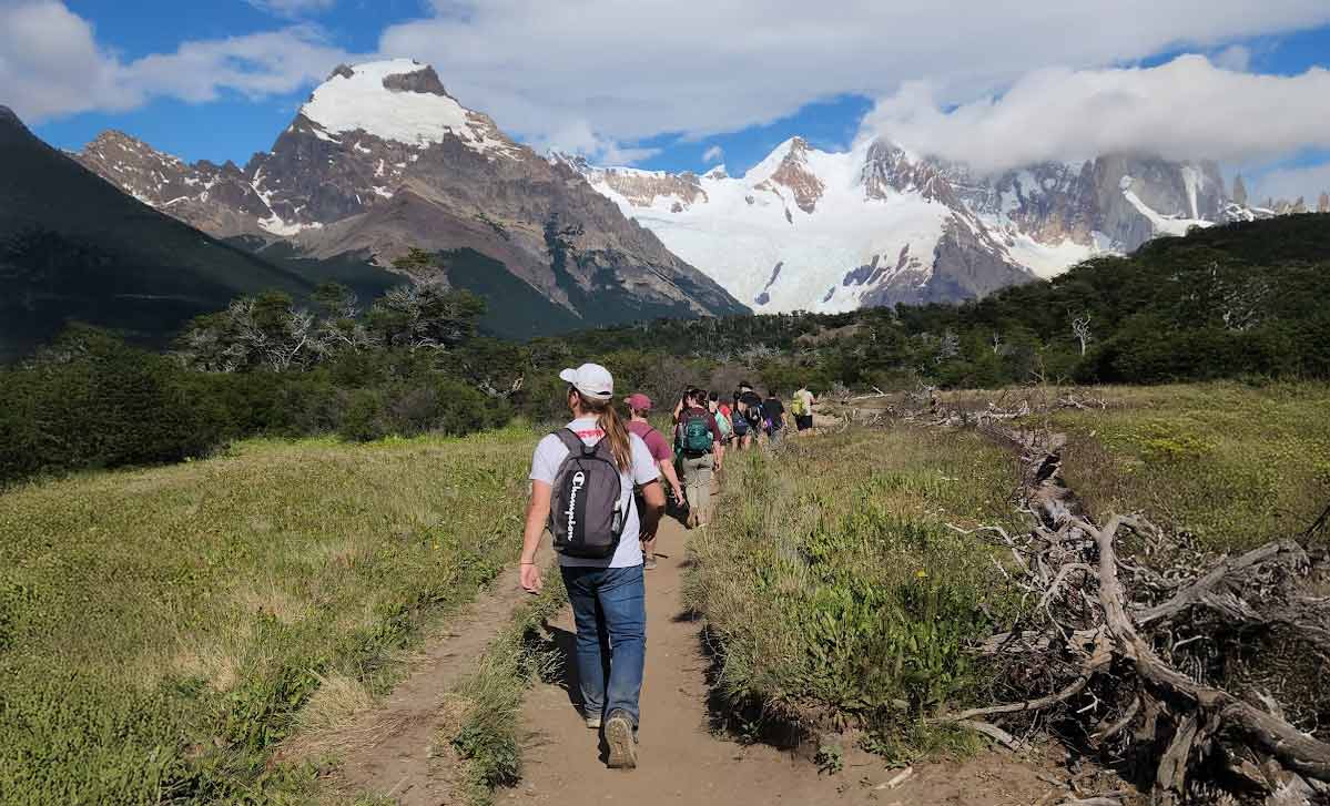 A group of Miami University students hiking the "Sendero a la Laguna Torre" Trail in El Chaltén, Argentina along a dirt trail through a green valley toward snow-covered mountains under a partly cloudy sky.