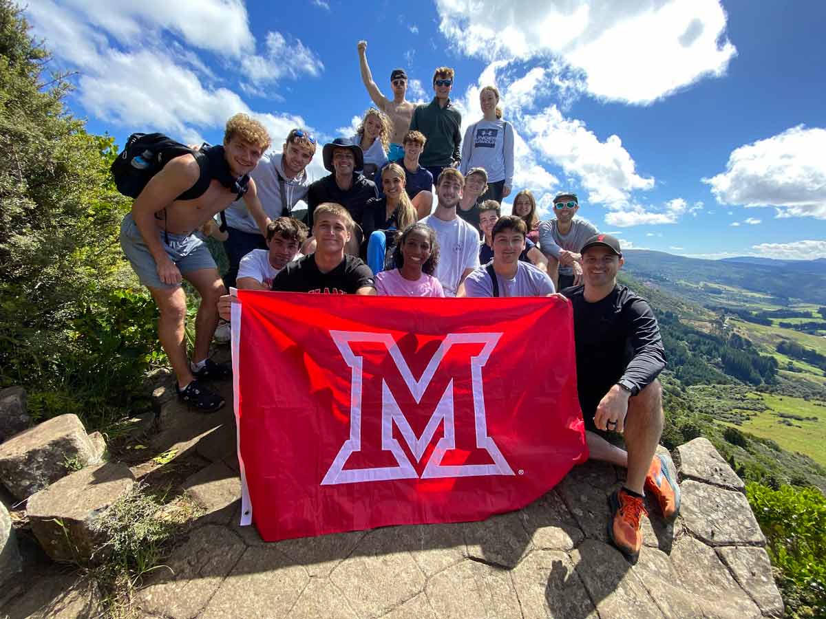 A group of Miami University students pose at a scenic mountain summit, holding a red Miami “M” flag.