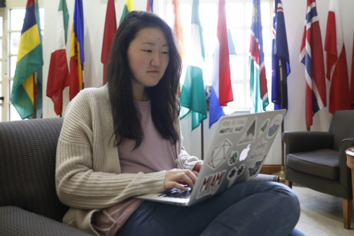 A student looking for information on her computer while sitting in the lobby of MacMillan hall. The walls are lined with the flags of many countries.