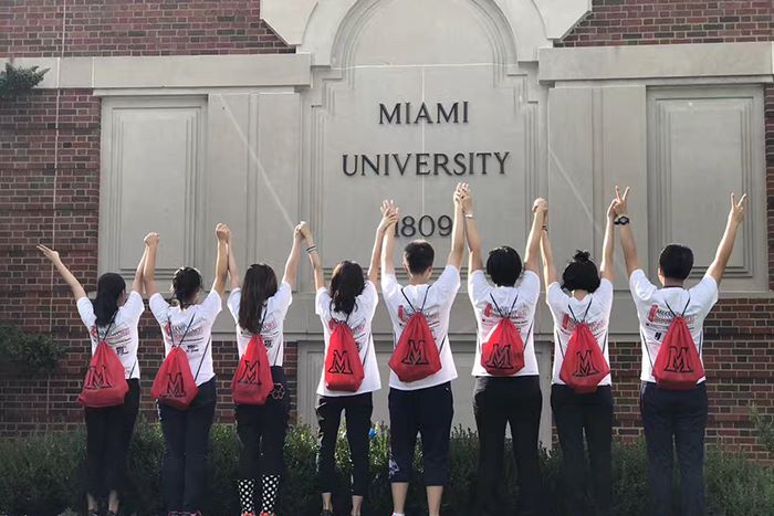 Students wearing matching Miami backpacks pose with their arms in the air in front of a large Miami University sign