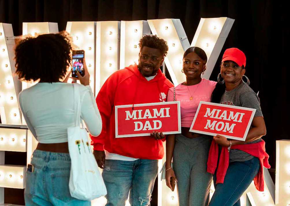 A student poses with her parents holding “Miami Dad” and “Miami Mom” signs as another student takes their photo in front of large lit-up letters spelling out FAMILY WEEKEND.