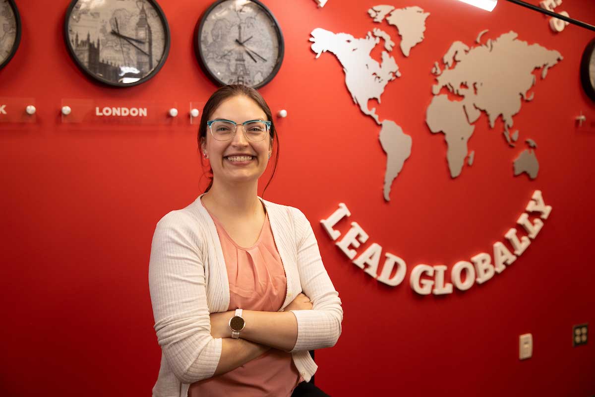 A smiling student stands in front of a red wall with clocks labeled by global cities and a silver world map installation that reads “Lead Globally”.