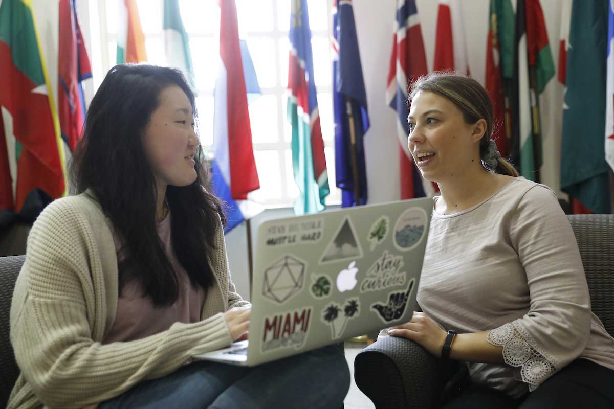 Two students talk while sitting in front of a display of international flags.